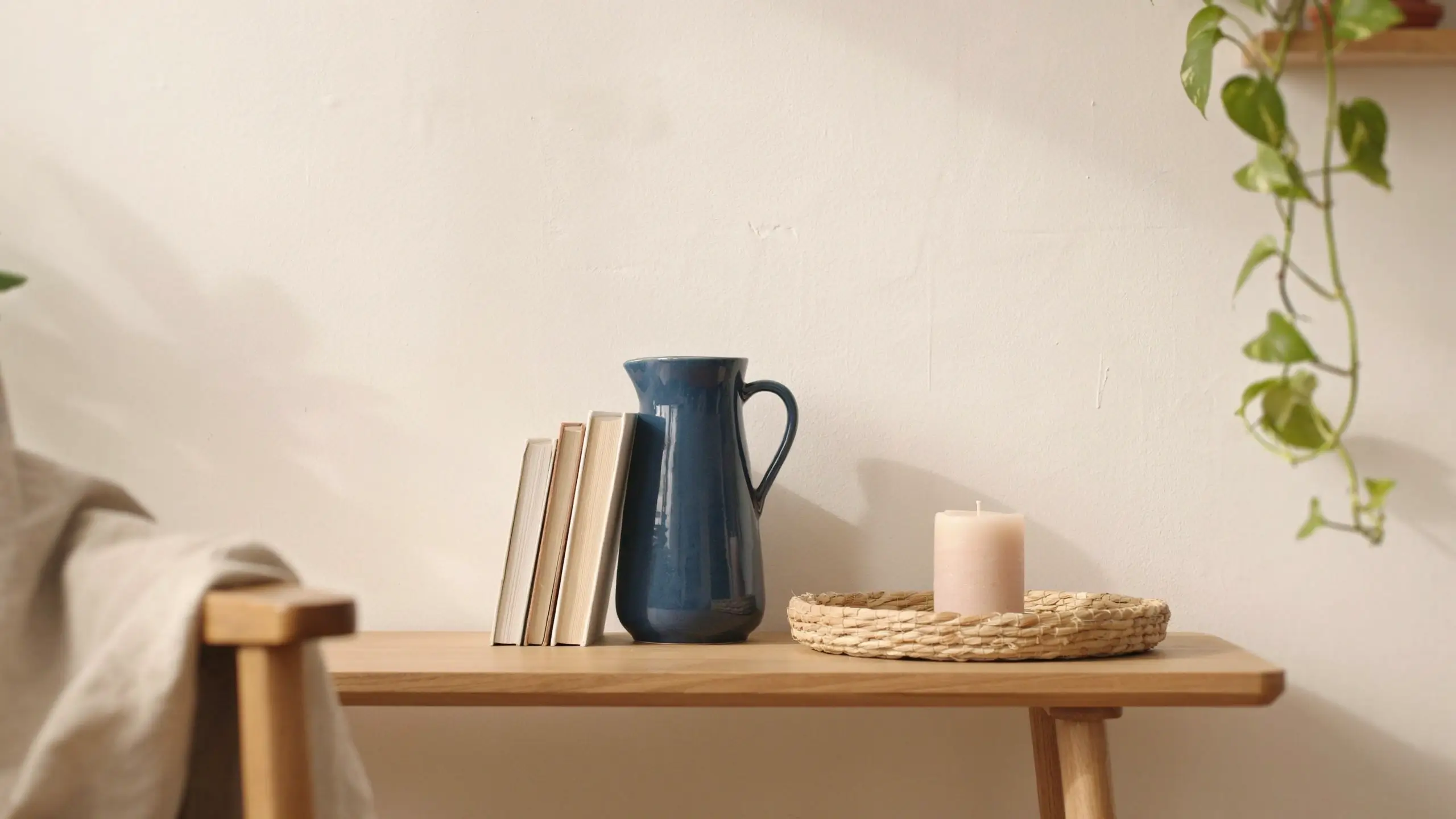 A minimalist neutral living room arrangement on a light wooden table featuring a deep blue ceramic pitcher and stack of beige books on the left, alongside a cream pillar candle on a woven rattan tray, with a trailing green pothos plant and a beige linen throw to demonstrate using accessories to shift the color palette.