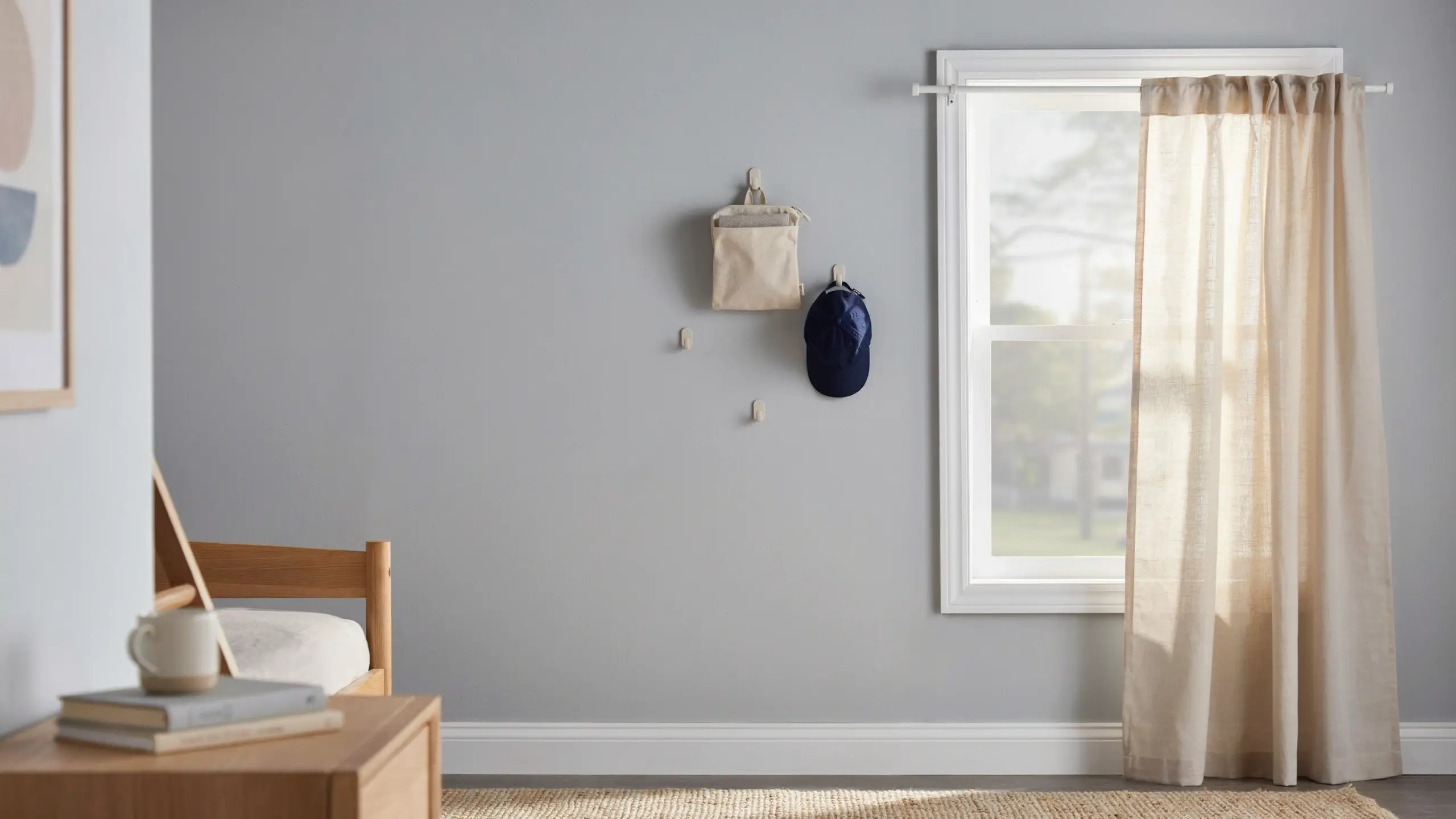 Beige command hooks holding a canvas pouch and blue cap, paired with a white tension rod and sheer curtain for damage-free wall storage in a sunlit rental space.