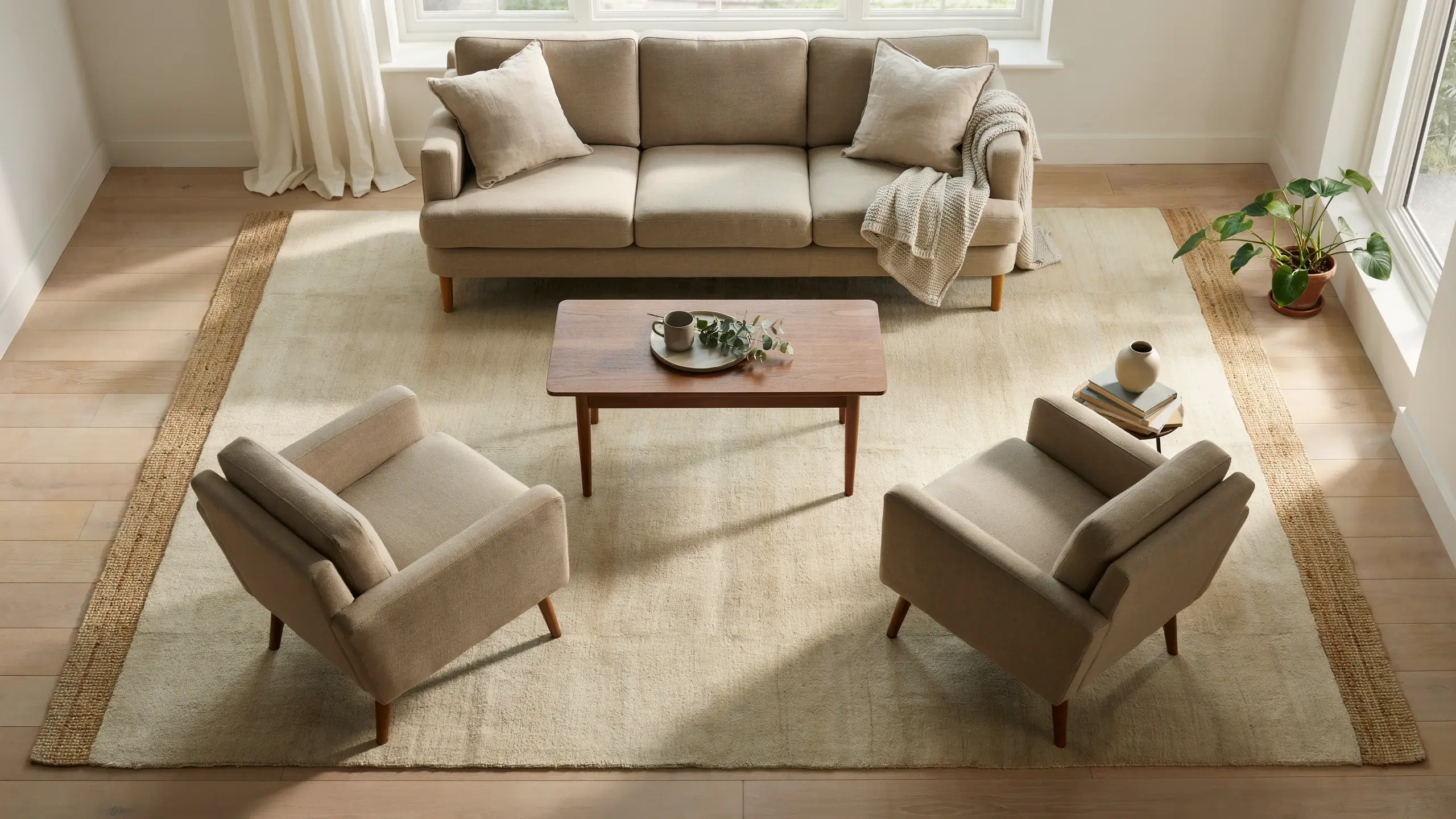 A modern living room featuring a large beige area rug with a textured brown border placed under a matching beige sofa and two armchairs, demonstrating the ideal rug size and proper furniture placement for anchoring the seating area.