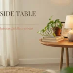Round wooden bedside table styled with a ceramic lamp, stacked books, and a potted pothos plant next to a cozy bed. Text overlay reads: 21 bedside table ideas that make your bedroom feel like a retreat.
