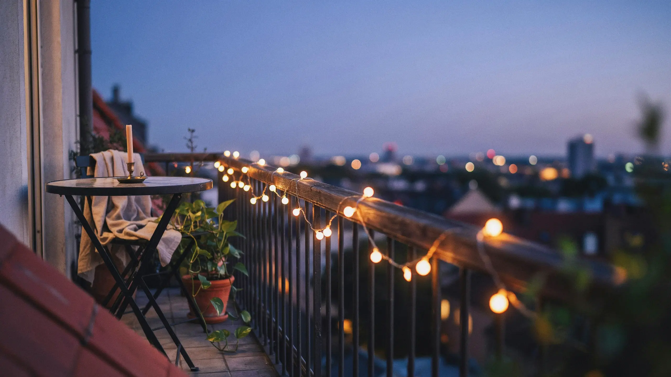 A cozy evening balcony featuring warm string lights draped along the railing, a small table with a throw blanket and candle, potted plant, and a blurred city skyline.