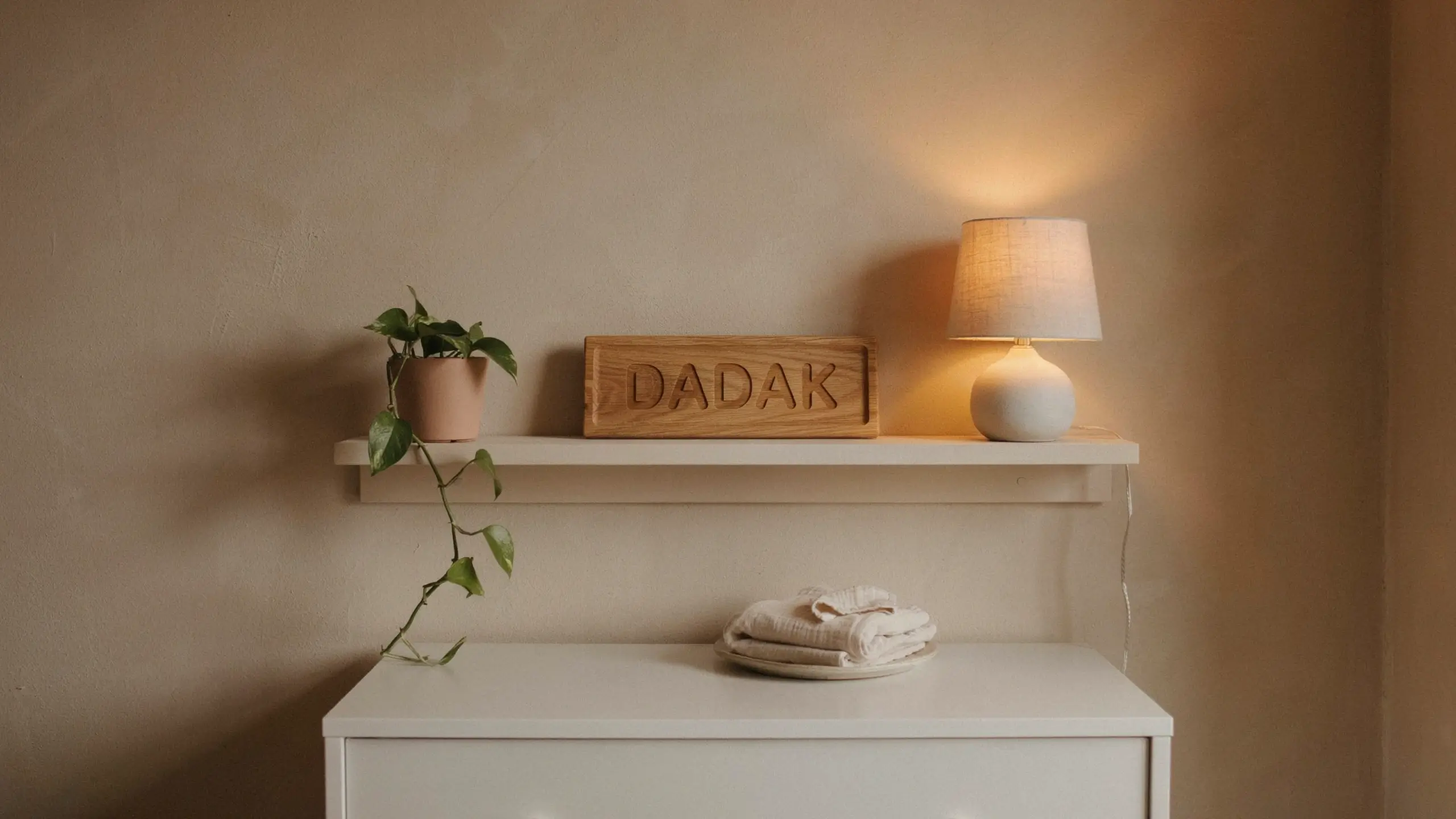 White floating shelf holding a terracotta potted pothos plant, a rectangular light wood sign with the carved text "DADAK", and a glowing white ceramic table lamp with a beige fabric shade, set against a warm beige wall above a white cabinet holding a stack of folded white clothes.