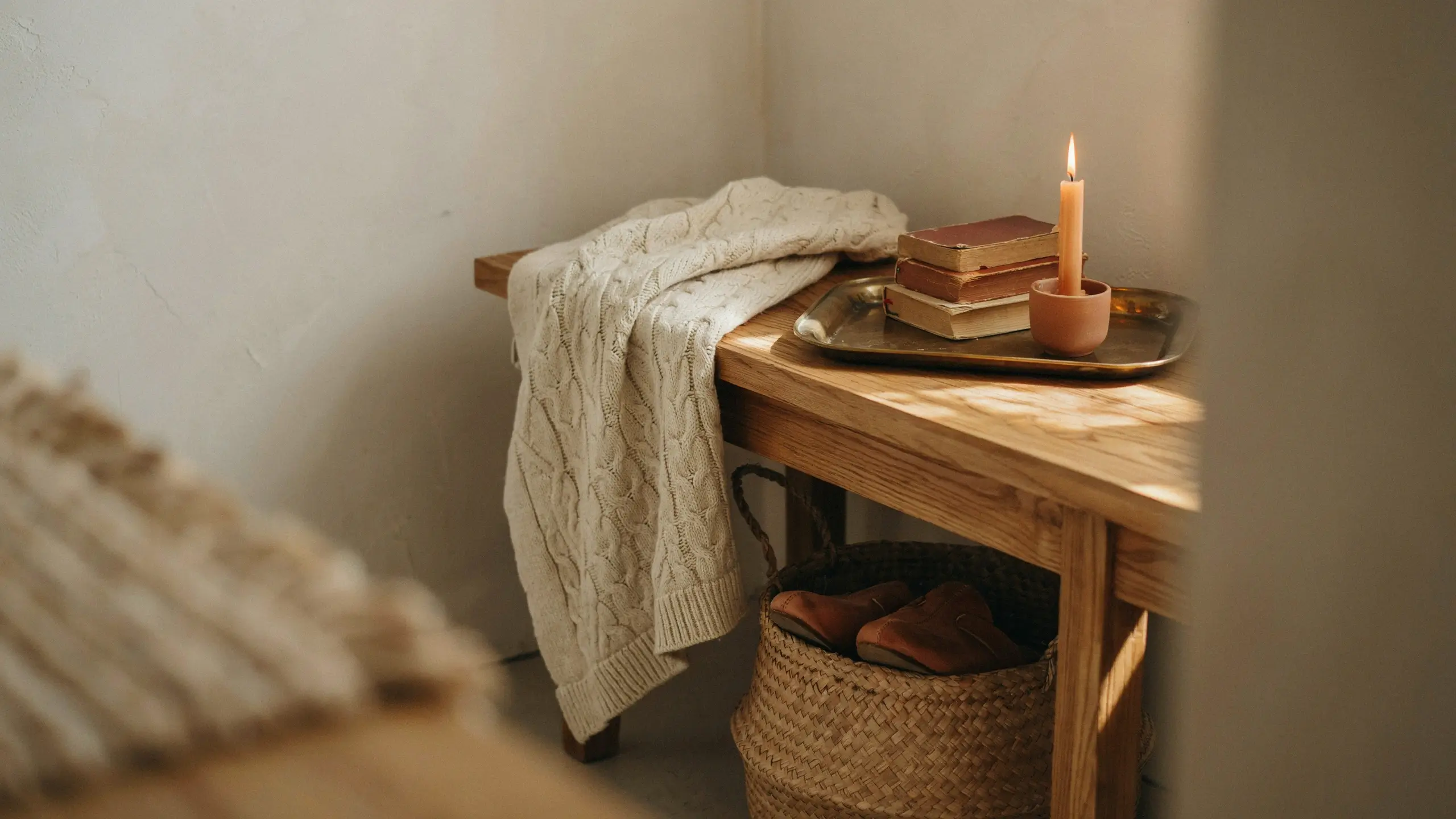 A warm, minimalist corner featuring a rustic wooden bench draped with a cream-colored chunky knit sweater. A vintage metal tray holds two stacked books and a glowing candle in a terracotta holder, enhancing the cozy personality of the space.