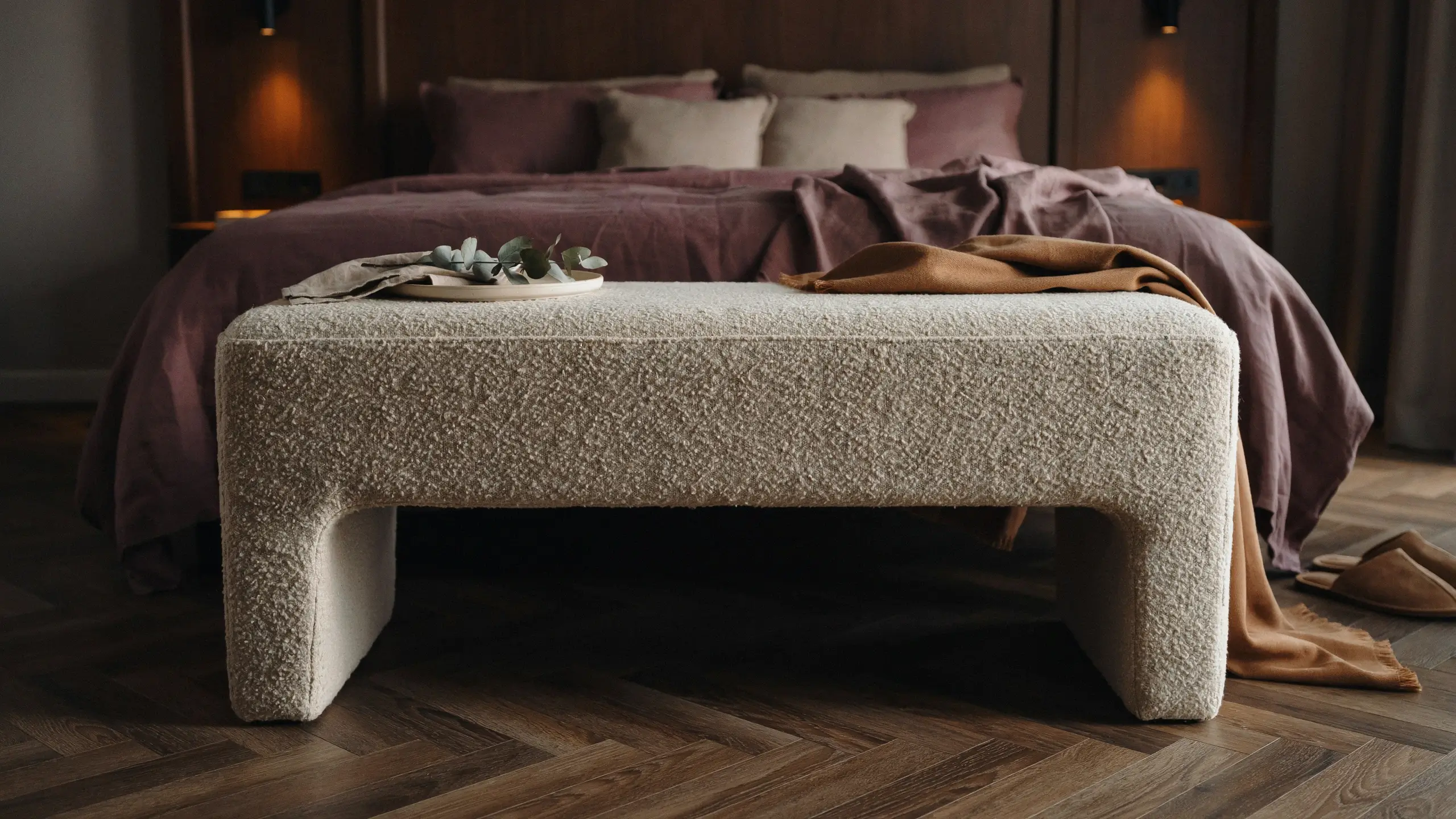 Cream textured bouclé bench at the foot of a bed with dusty rose linens, a draped tan throw blanket, and a tray with eucalyptus leaves in a cozy bedroom.