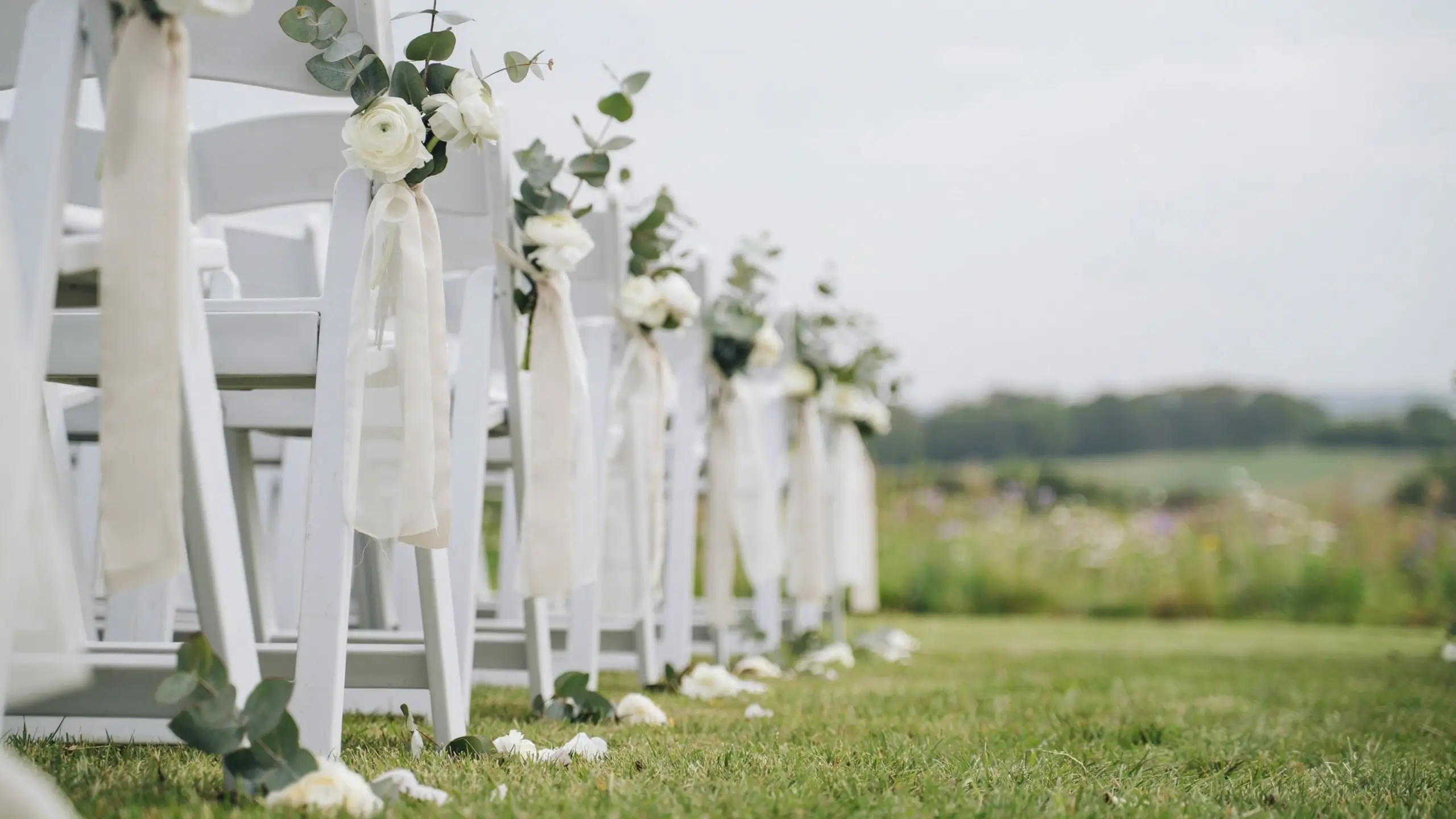 White wooden wedding chairs set in a garden aisle, adorned with white flowers, eucalyptus, and flowing curtain panels tied to serve as rustic aisle markers.