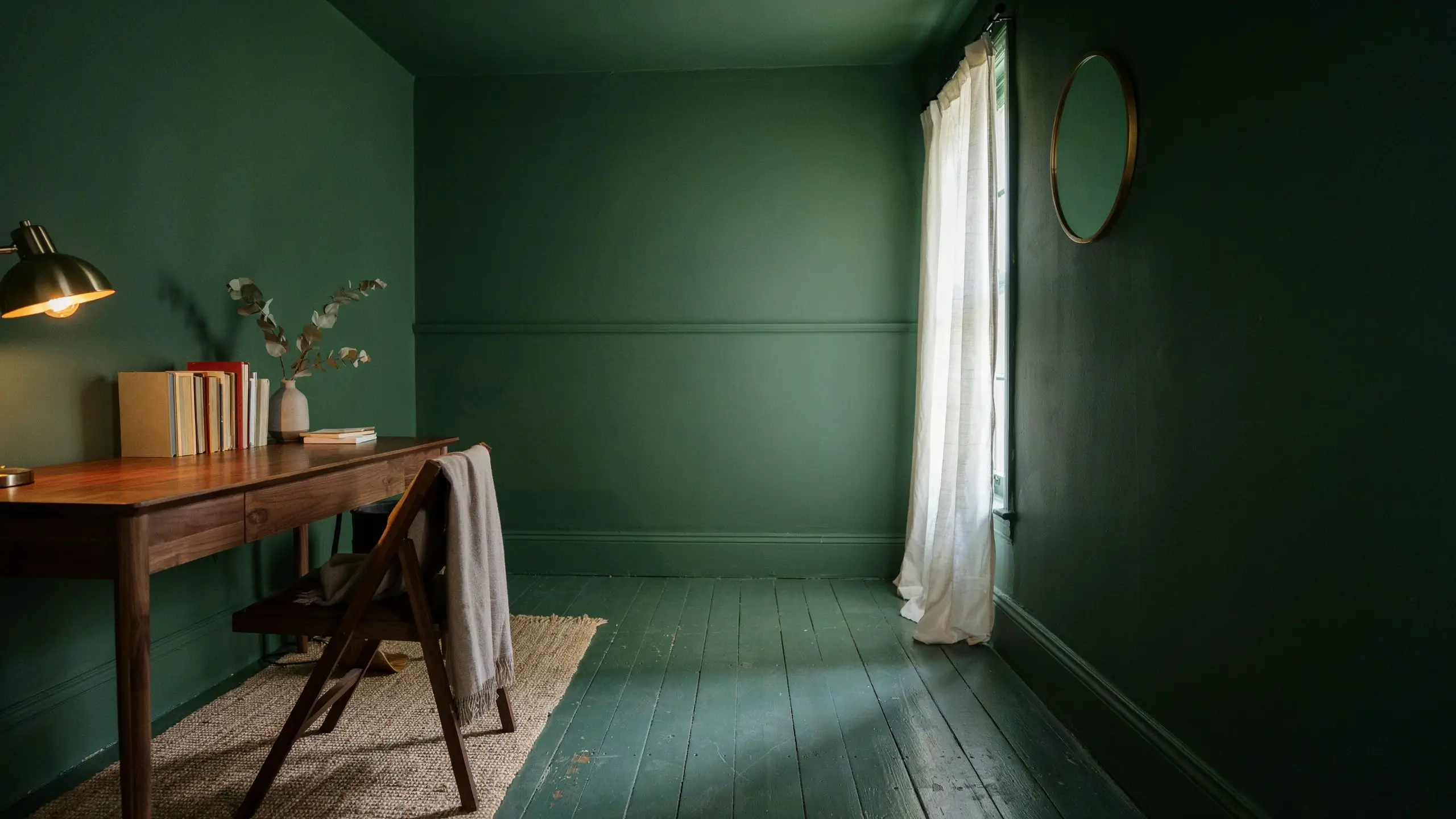 A moody, dark green small office nook featuring sheer white curtains, a round brass mirror, and a wood desk that uses natural window light to visually expand the space.