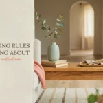 A bright, minimalist living room featuring a wooden coffee table styled with a sage green vase holding eucalyptus, a stack of books, and a wooden tray with dried grass. A white sofa with a pink throw blanket sits beside a white wall with text overlay reading '9 Decorating Rules I Was Wrong About', serving as a cover image for an interior design blog post.