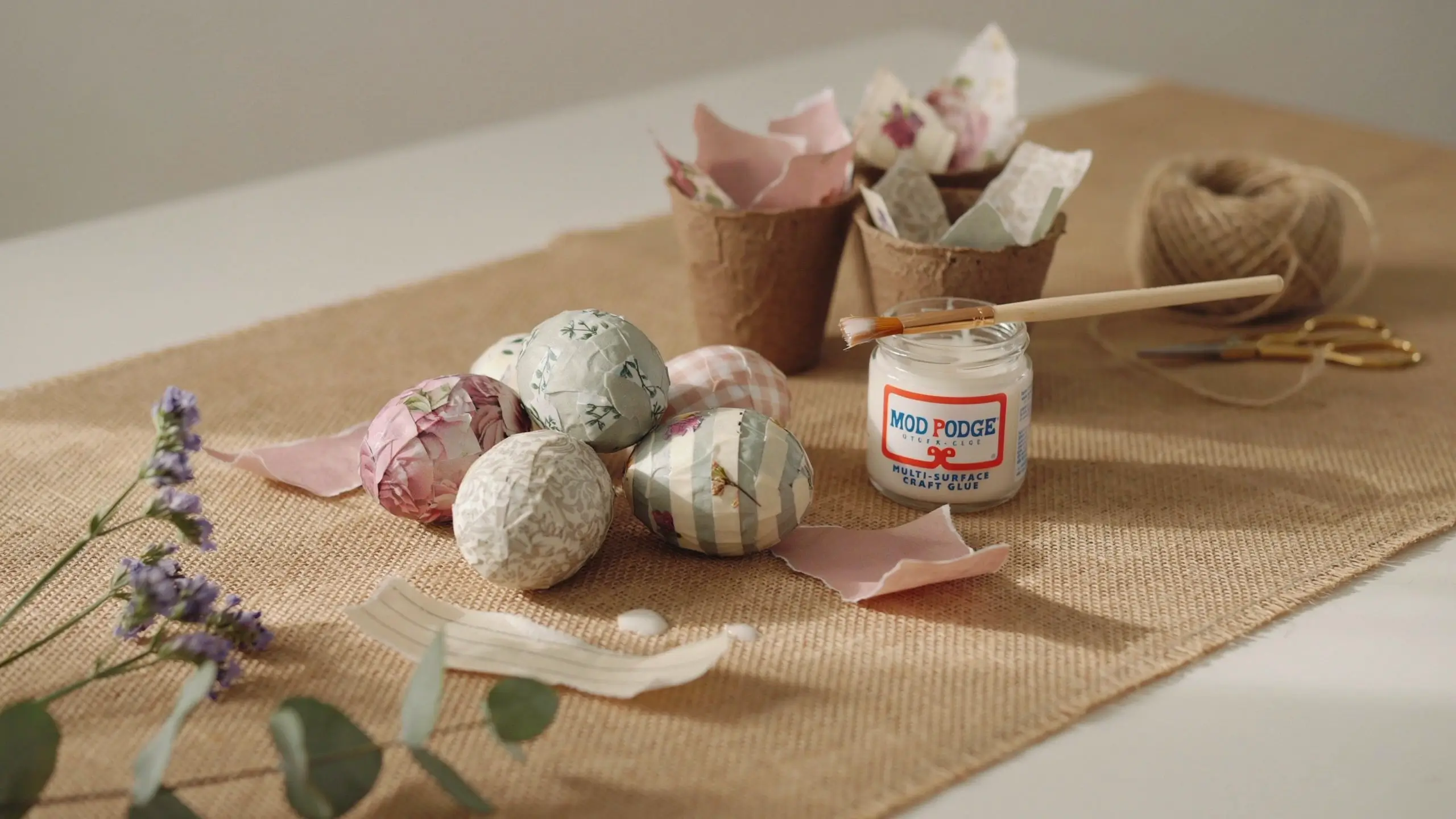 DIY decoupage Easter eggs featuring floral and striped patterned paper resting inside rustic peat pots, with a jar of Mod Podge and a wooden brush on a burlap craft mat.