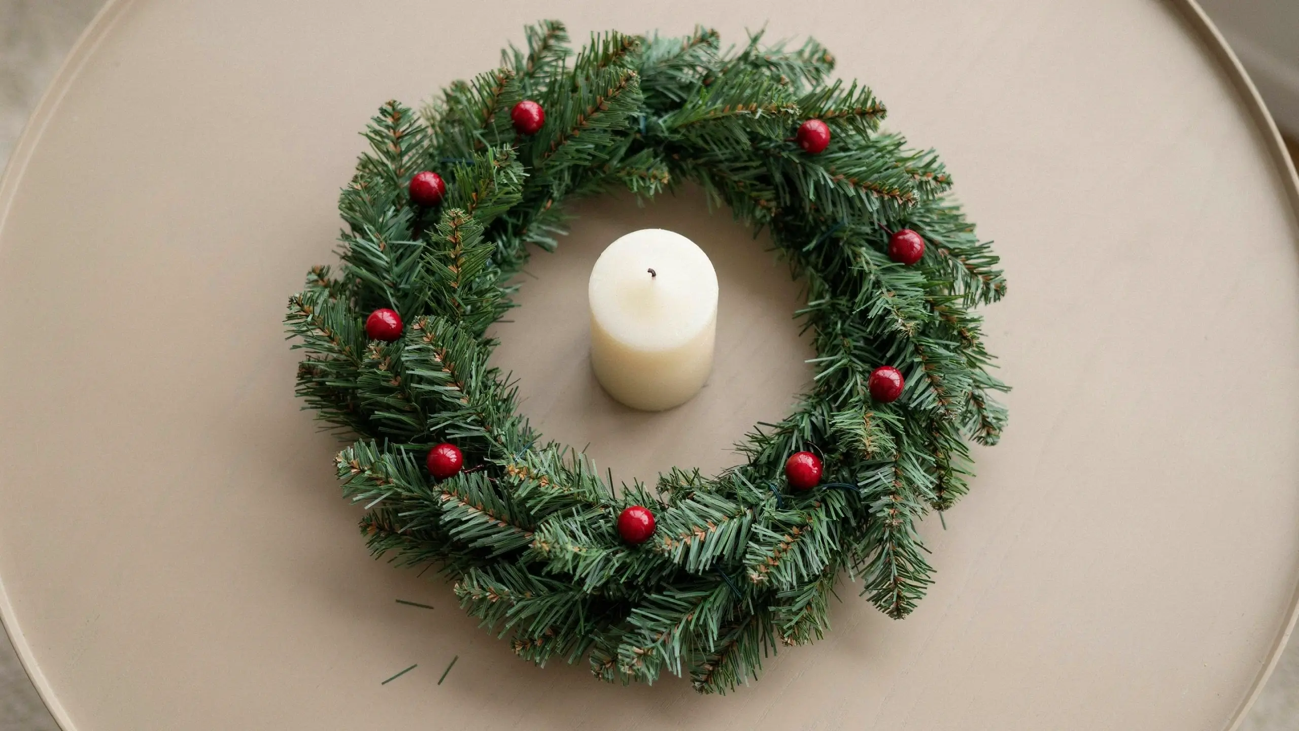 Minimalist DIY Christmas wreath centerpiece made from a coiled dollar store pine garland, accented with bright red berries and a white pillar candle on a neutral beige round table.