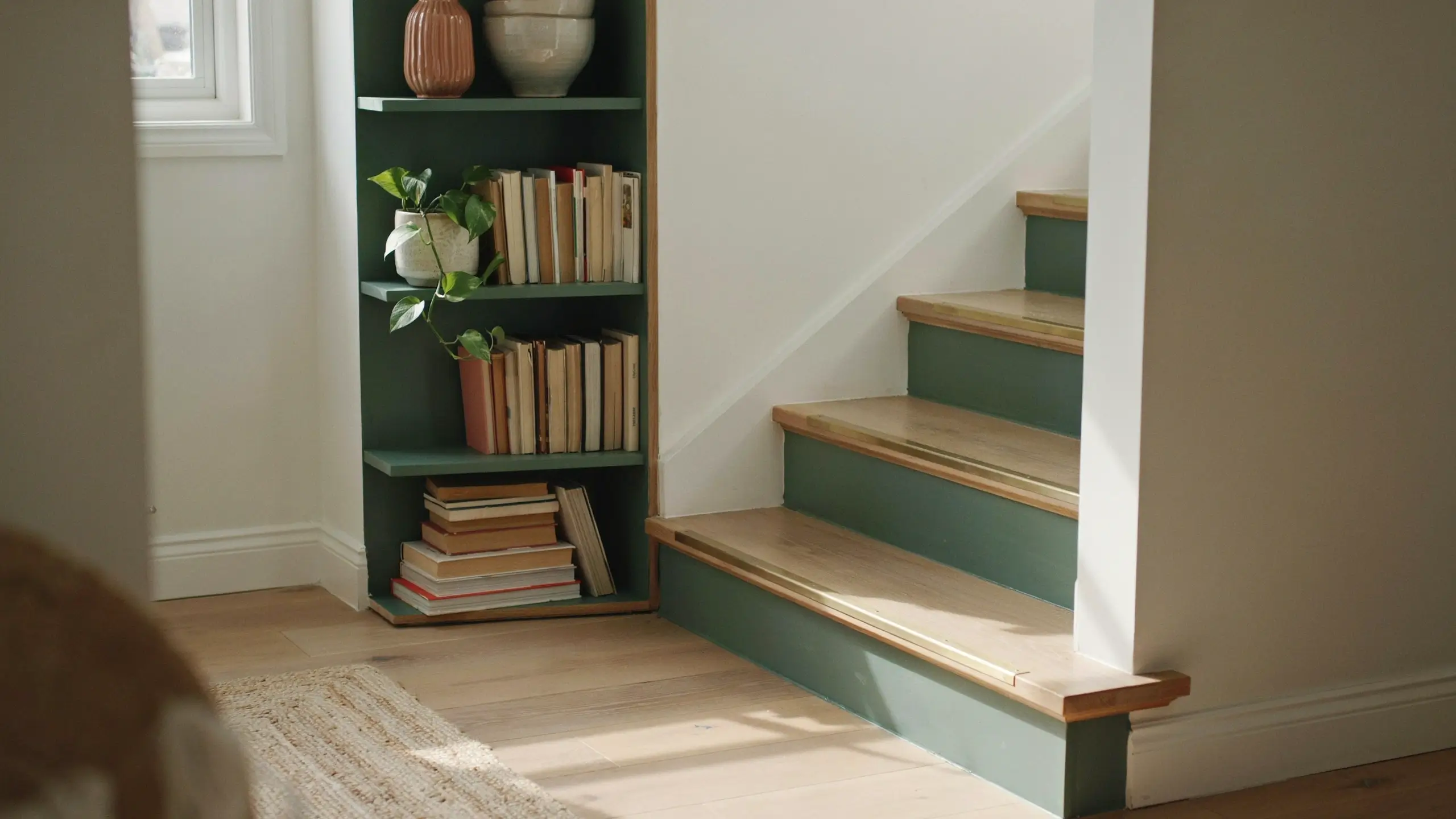 A built-in green bookshelf filled with books and a trailing pothos plant, located right next to a staircase featuring DIY painted green risers and natural wood treads