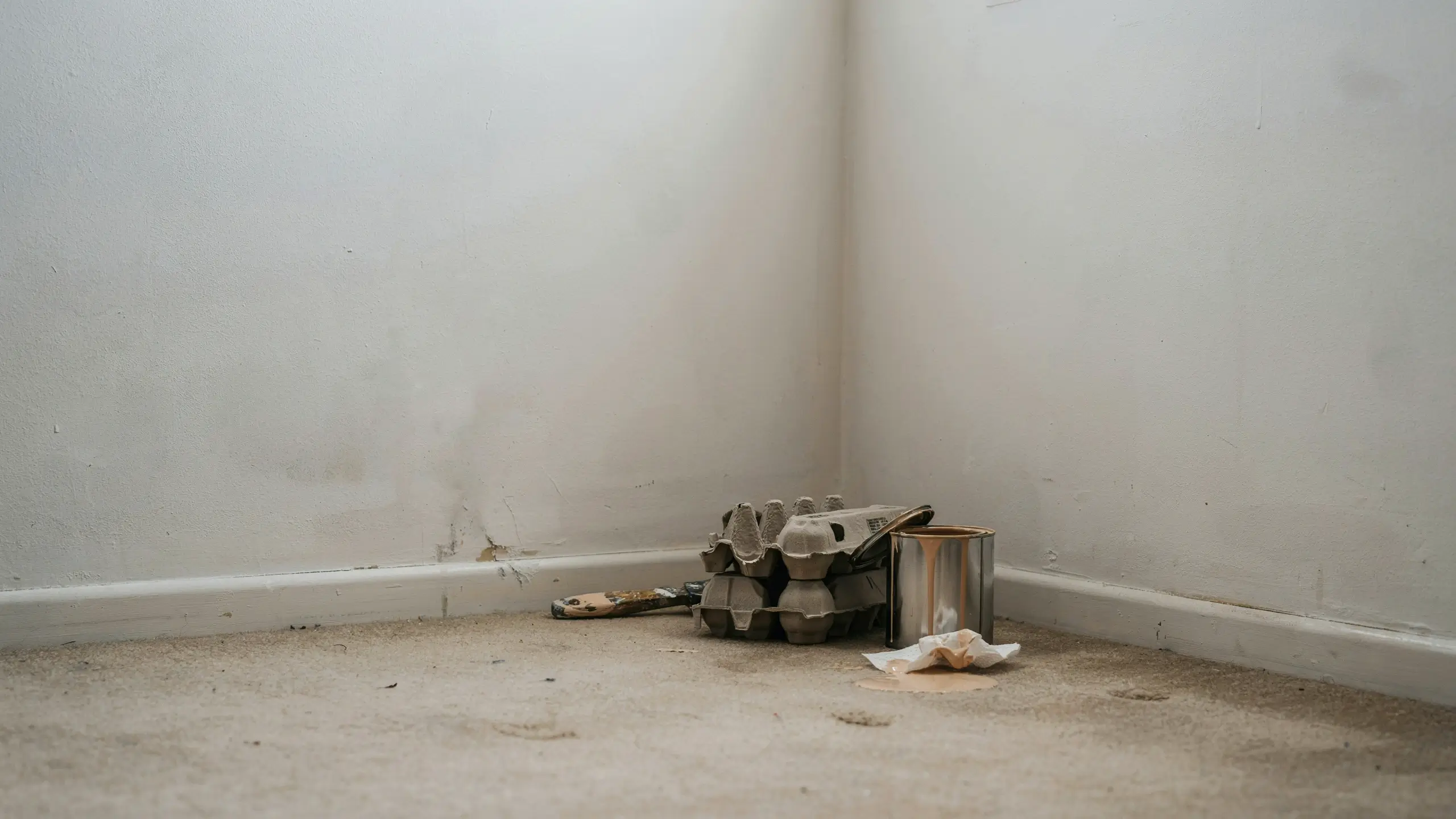 A messy corner of a freshly painted room showing used DIY supplies, including a metal paint can, dirty brush, stained rag, and a cardboard egg carton on a dusty floor, illustrating common home renovation errors.