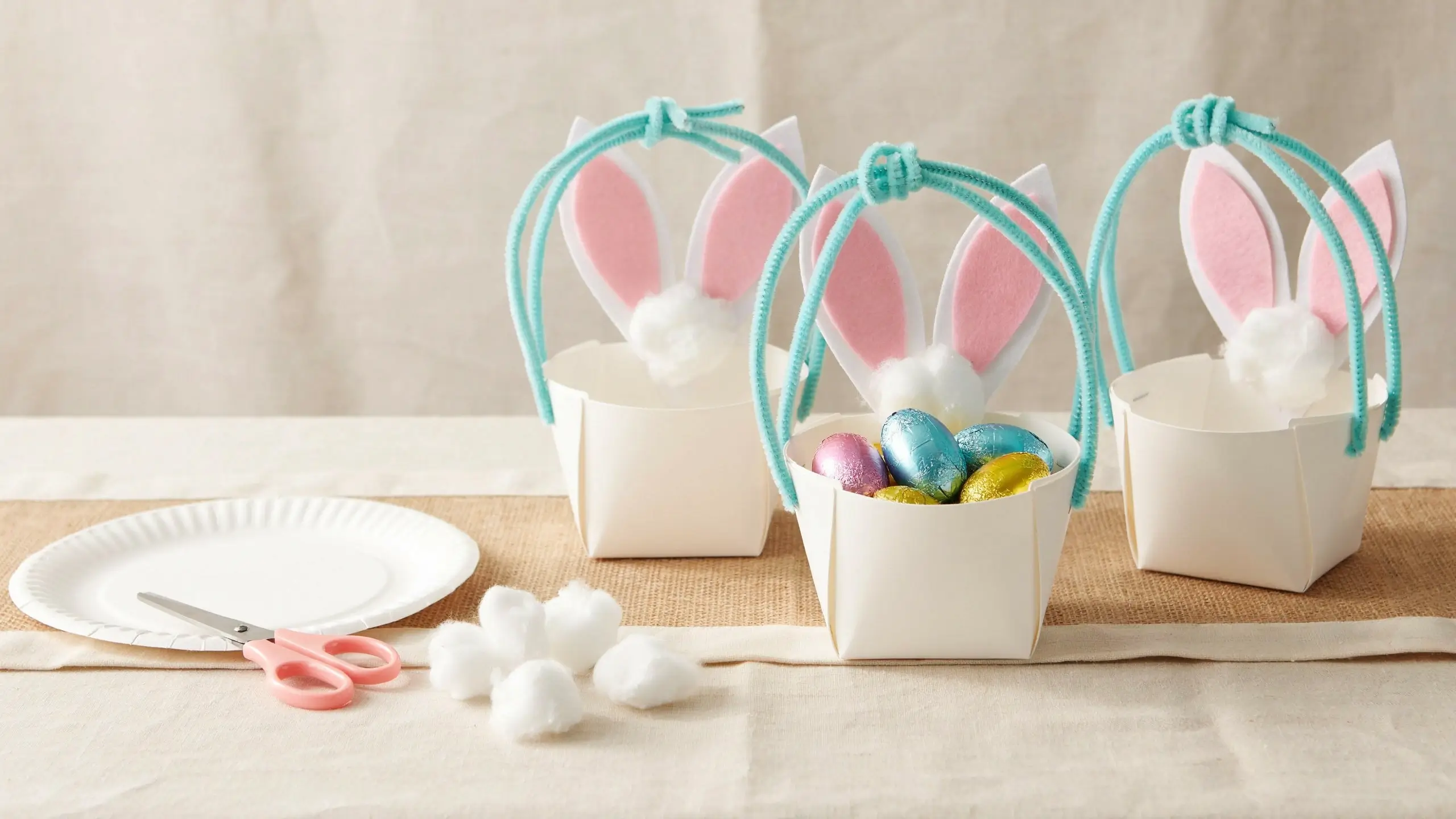 Three handmade paper plate Easter bunny baskets filled with colorful foil-wrapped chocolate eggs, featuring pink felt ears, blue pipe cleaner handles, and white cotton ball tails, surrounded by pink crafting scissors and cotton balls.