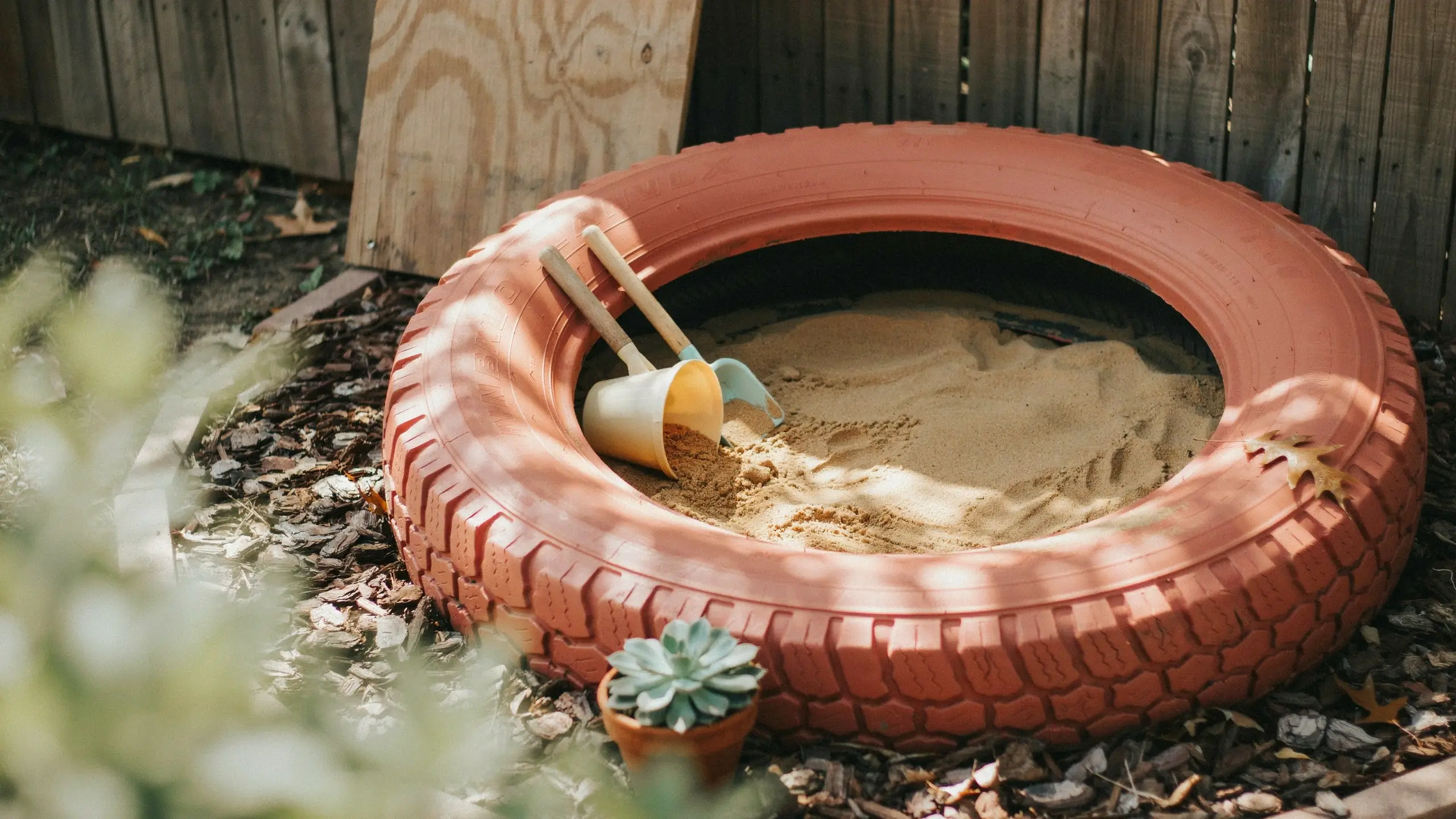 Orange repurposed tire sandbox filled with sand, toy bucket, and wooden shovels in a backyard setting