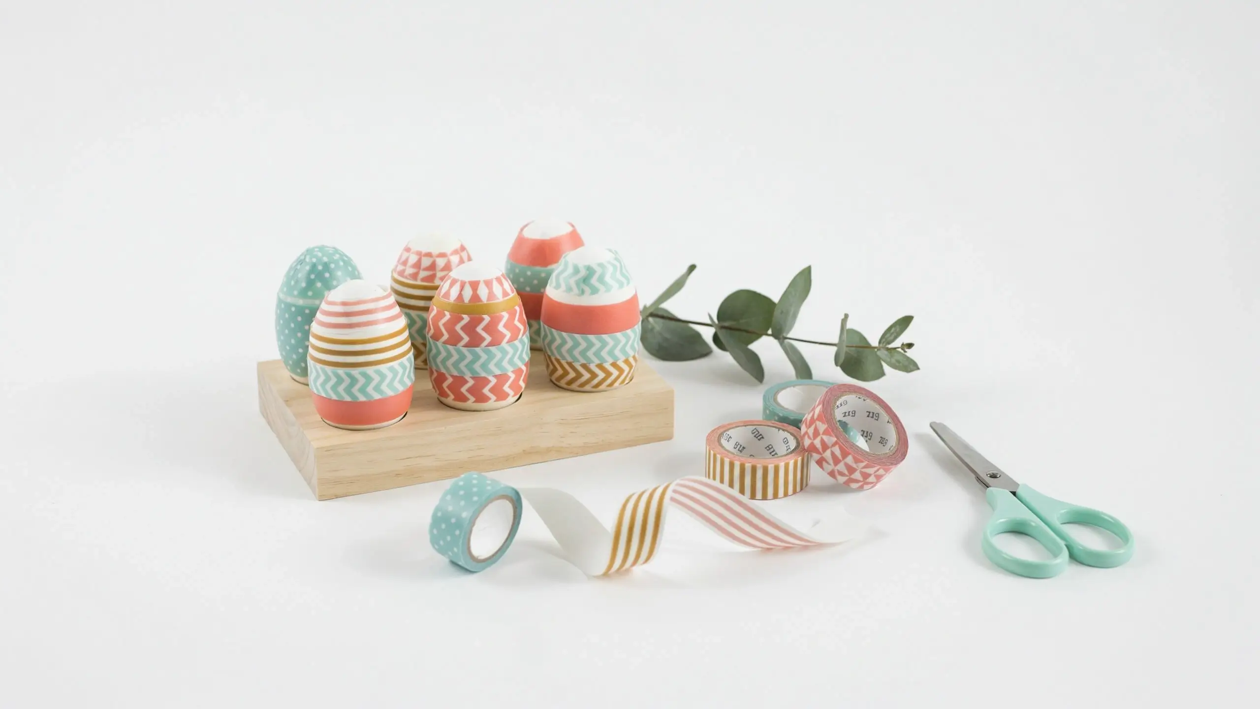 A minimalist flat lay of DIY washi tape Easter eggs in teal, coral, and gold geometric patterns resting on a small wooden stand, accompanied by matching patterned tape rolls, a mint green crafting scissors, and a sprig of fresh eucalyptus leaves on a white background.
