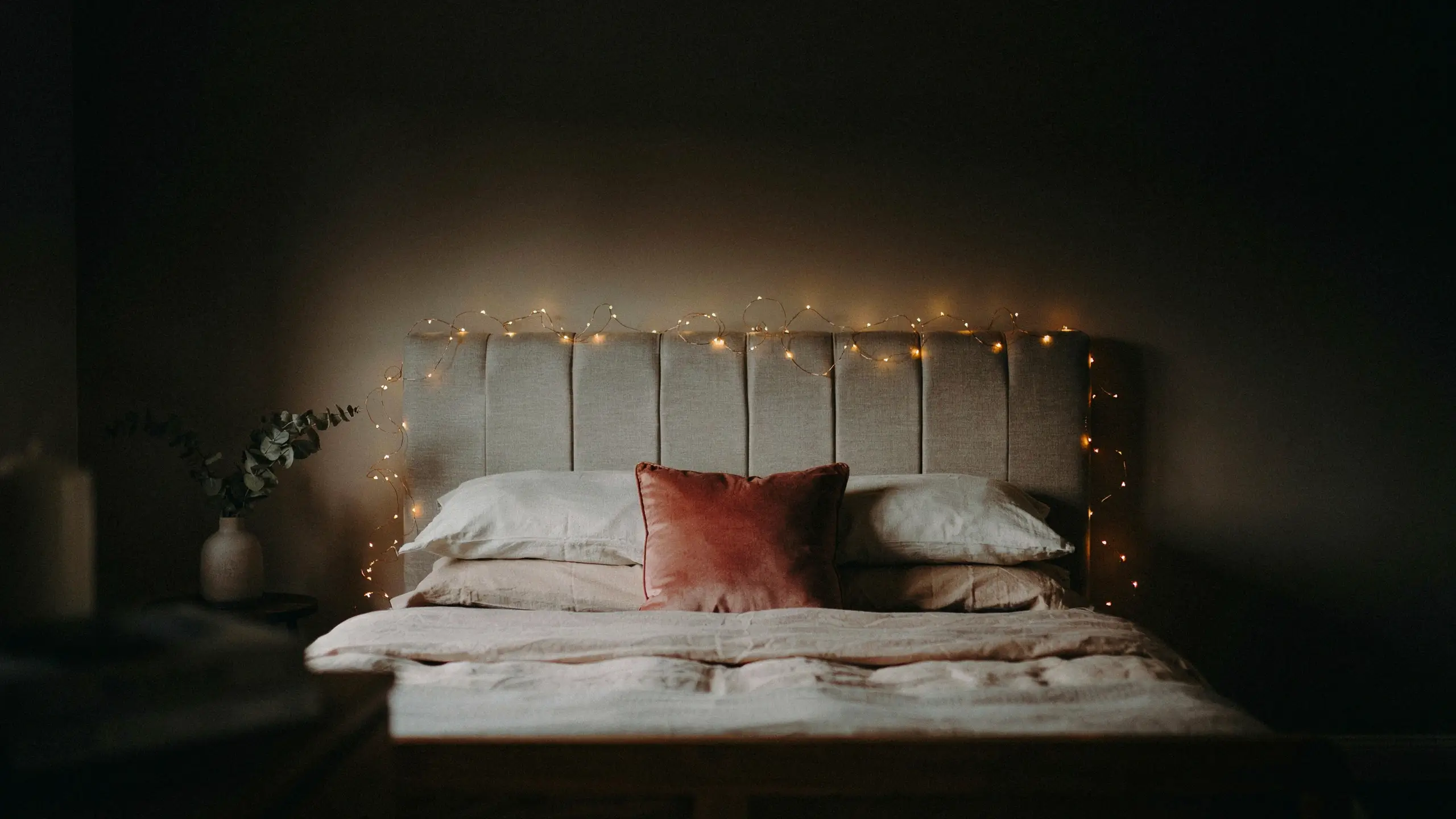 Beige upholstered headboard with warm string fairy lights draped across the top, featuring white pillows and a dusty rose velvet accent pillow in a dim, moody bedroom setting.