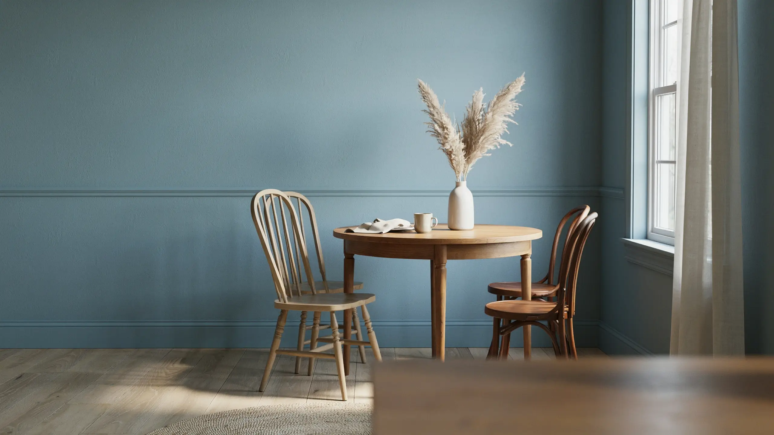 Bright dining room showcasing a monochromatic dusty blue wall and trim design with a round wooden table, white vase with pampas grass, and natural sunlight.