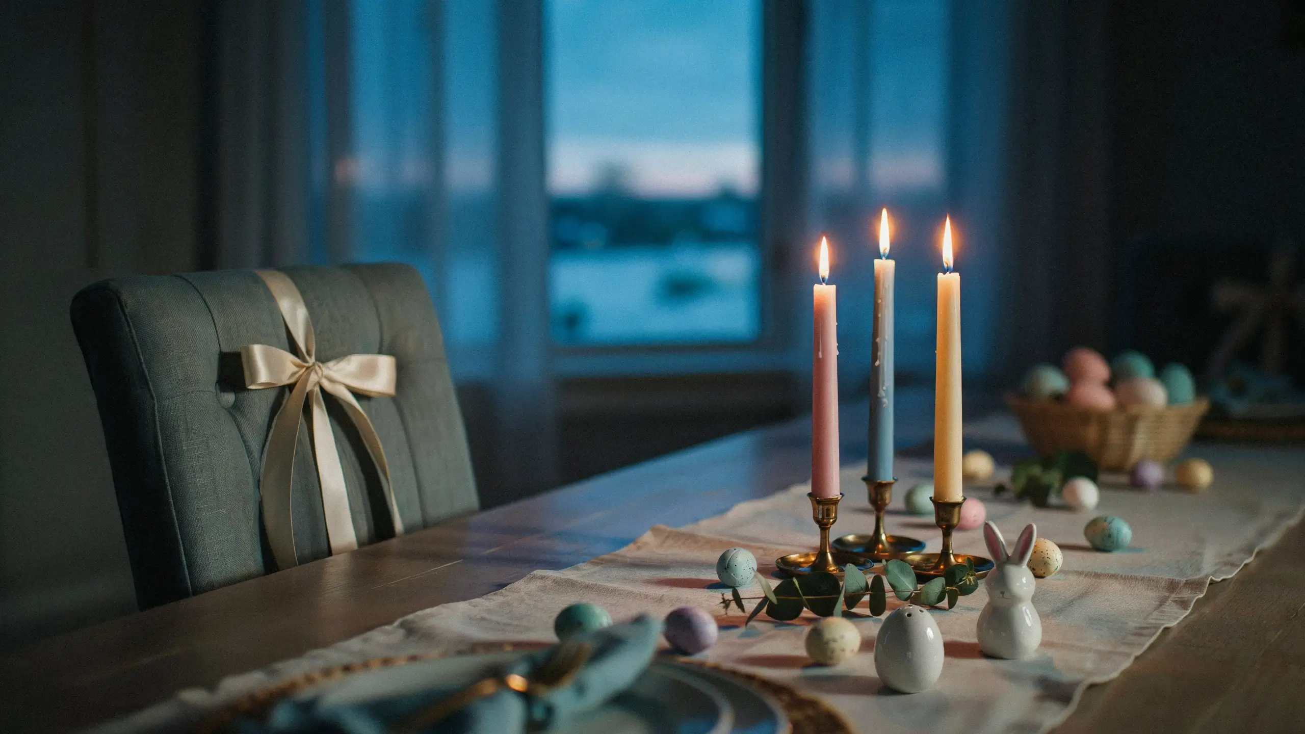 Easter candlelit table setup with pastel eggs, white bunny, and ribbon-adorned chair against a twilight window.