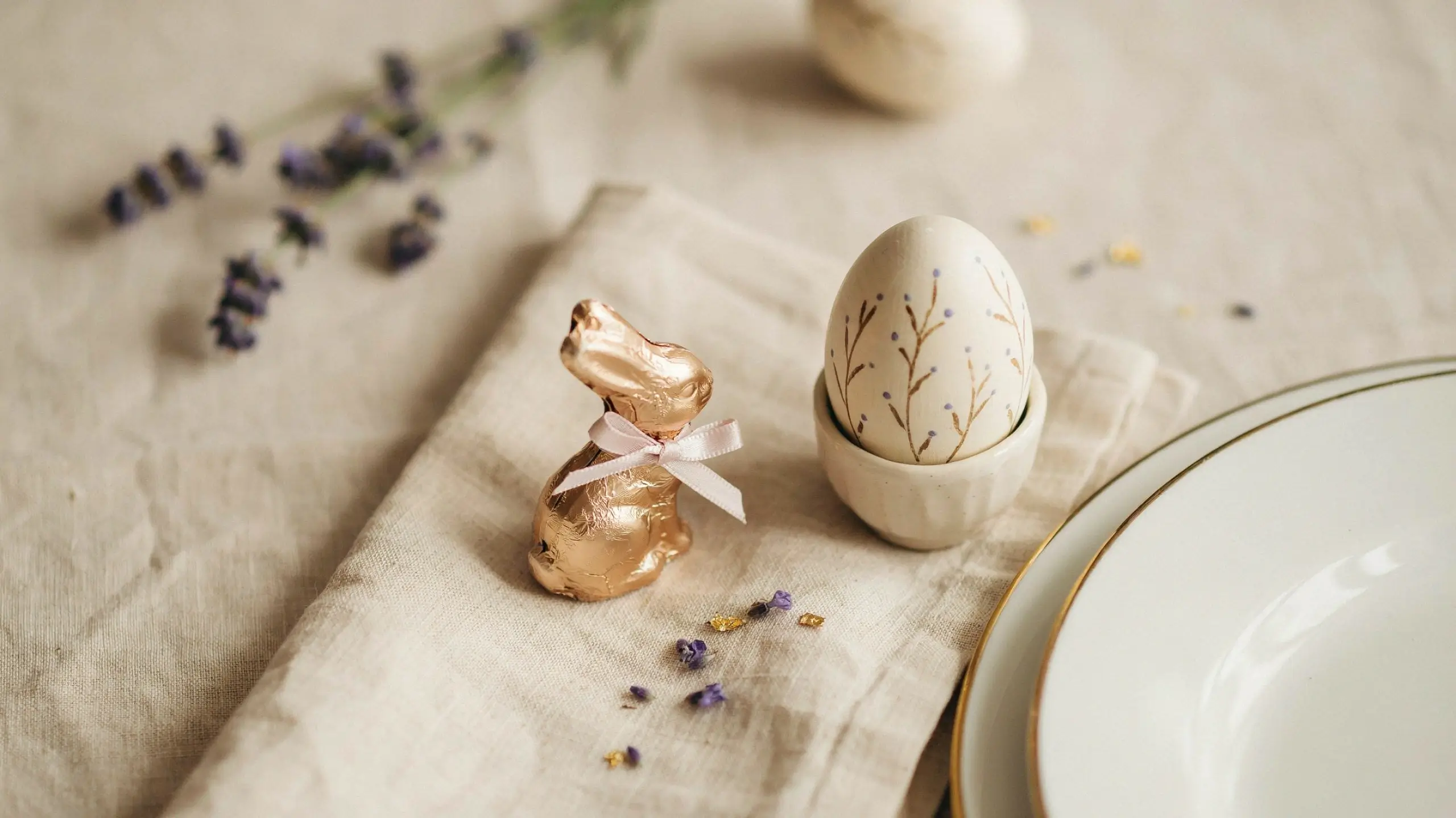 A minimalist Easter table setting with a gold foil chocolate bunny tied with a white ribbon and a botanical floral decorated egg in a ceramic egg cup, styled for elegant spring place cards.