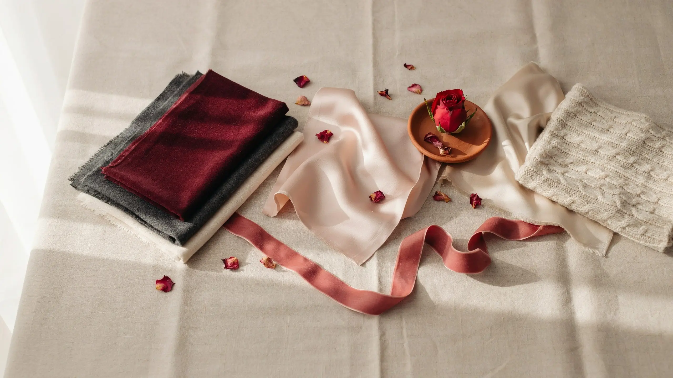 A soft, flat-lay arrangement of textiles featuring a folded burgundy scarf, grey wool, cream linen, draped blush pink silk, and a chunky cream knit sweater, scattered with red rose petals and a single pink rose on a terracotta dish, creating an effortless color palette.