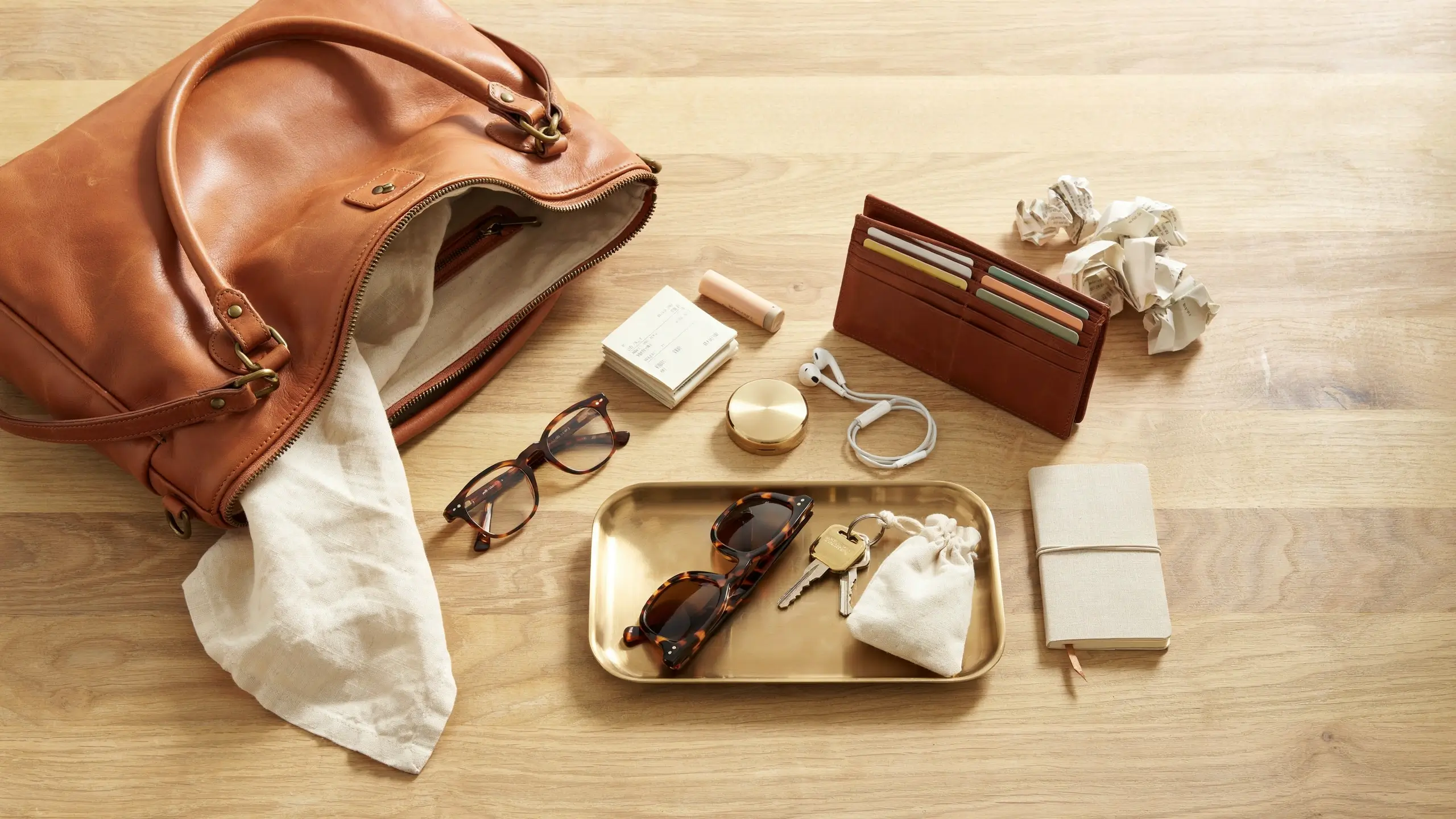 Open tan leather tote bag displaying a flat lay of everyday carry items including a brown card wallet, tortoiseshell sunglasses, car keys, white earbuds, and a notebook on a wooden surface.