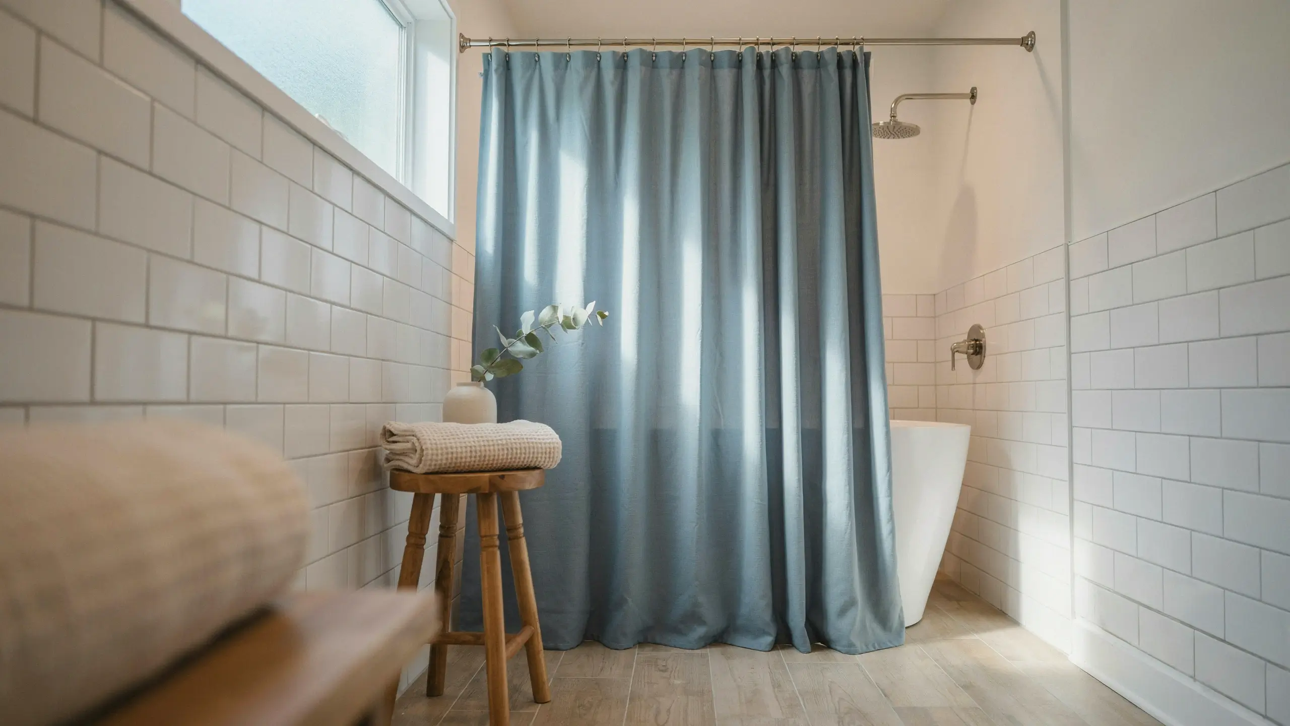 A serene bathroom featuring a slate-blue extra-long shower curtain hanging near the floor, alongside a wooden stool holding a folded towel and minimalist greenery, set against white subway tile walls and a freestanding bathtub.