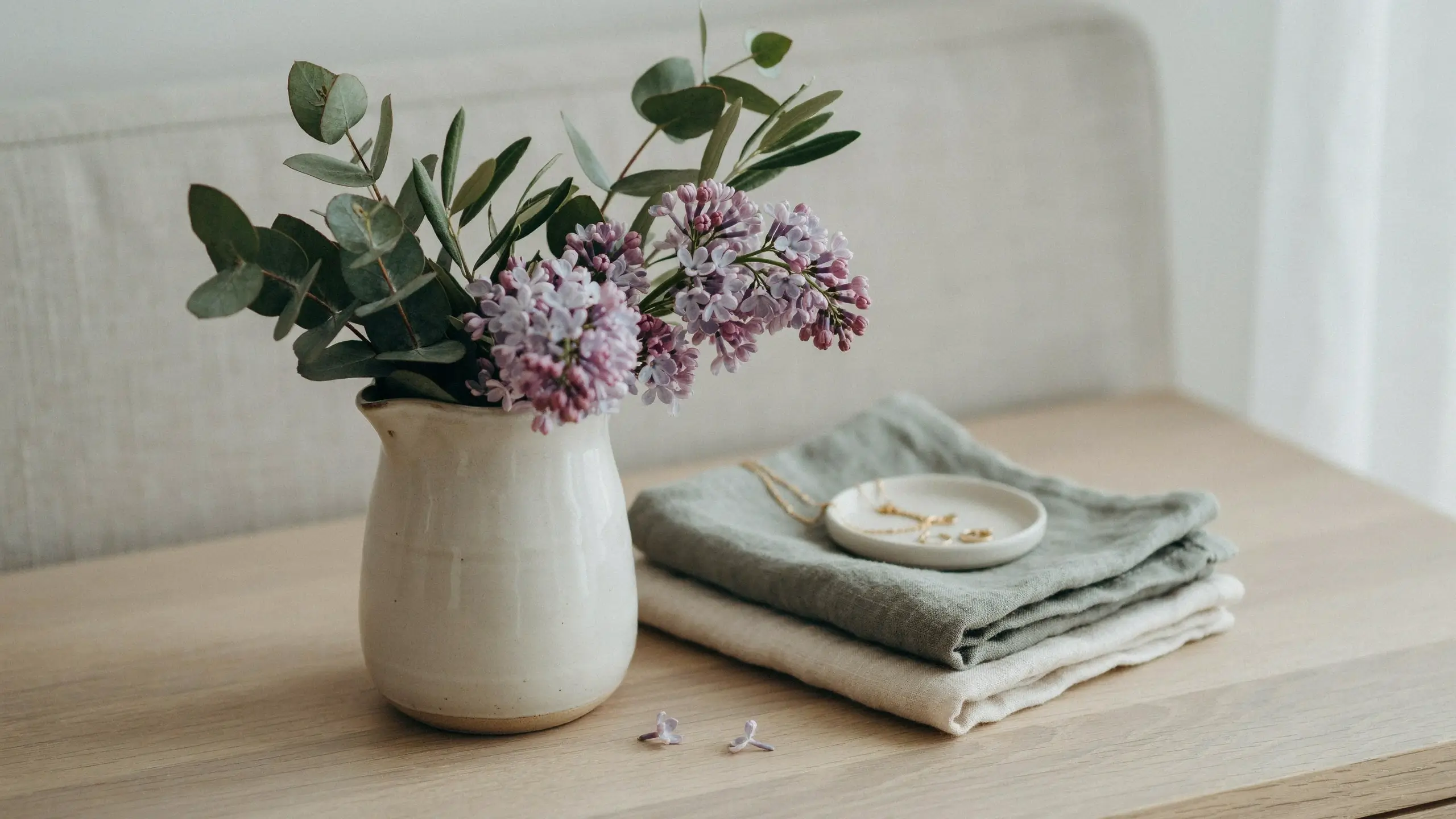 A cream ceramic pitcher filled with faux purple lilac branches and green eucalyptus leaves, resting on a wooden bedroom nightstand. Next to the vase is a stack of folded sage green and cream linen napkins topped with a white jewelry dish holding a gold chain, creating a serene and minimalist spring home decor aesthetic.
