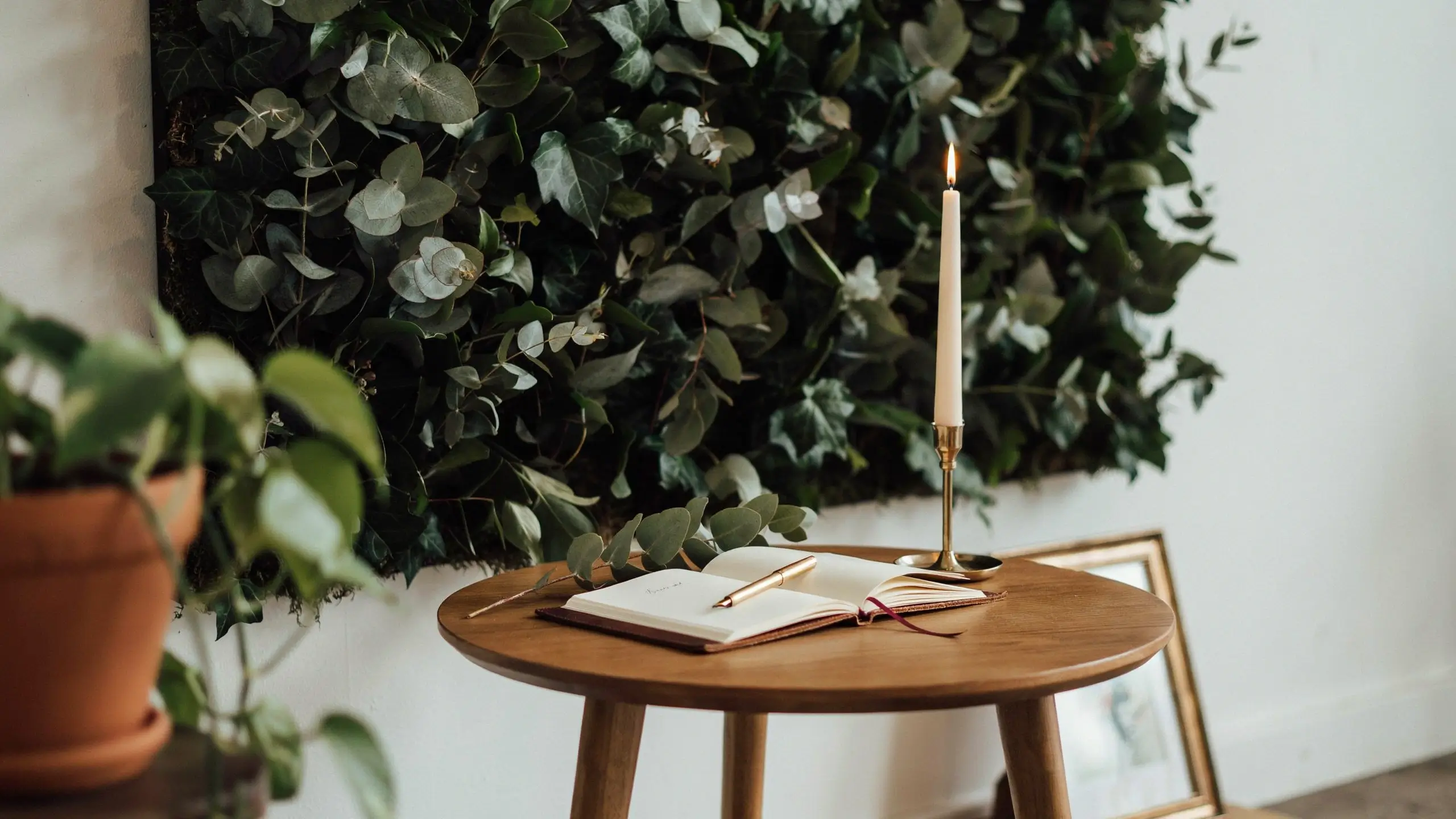 Close-up of a wooden table styled with an open guestbook, gold pen, and burning candle, positioned against a lush FEJKA artificial plant wall backdrop.