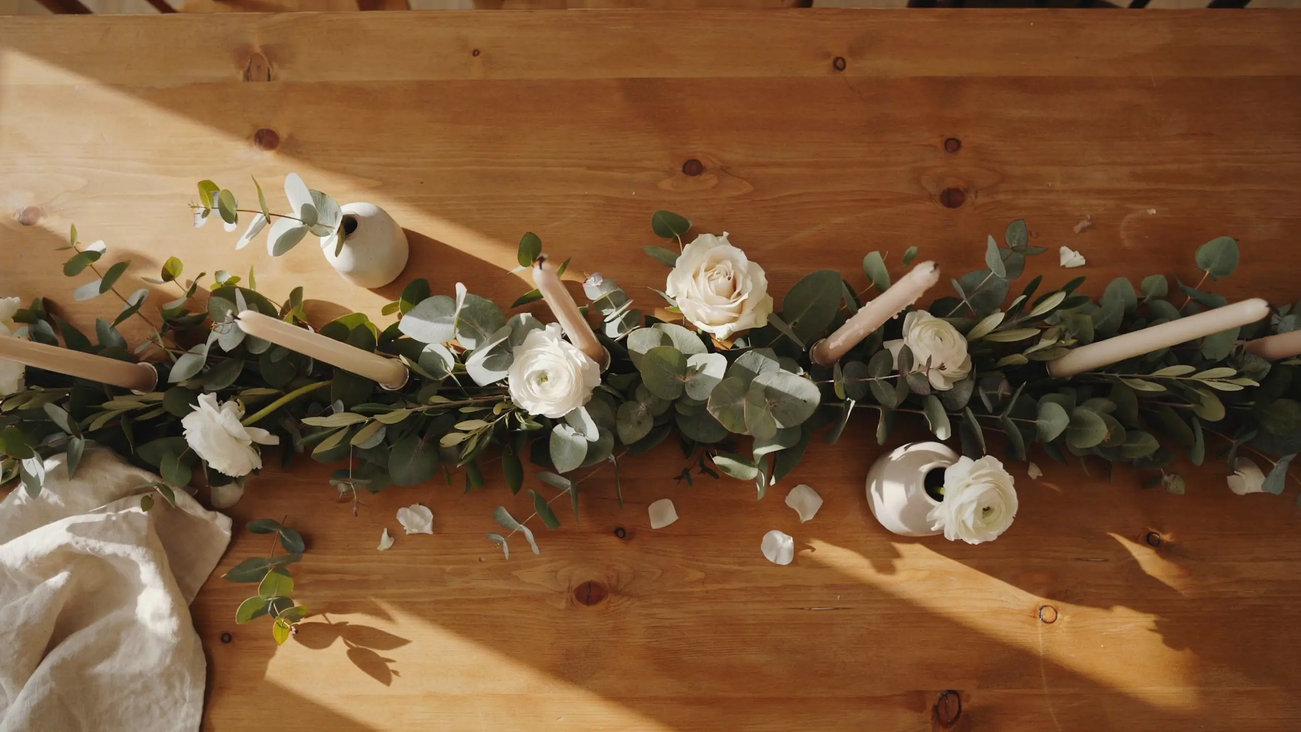 A top-down view of an artificial FEJKA eucalyptus and white rose garland styled as a table runner, featuring white taper candles, decorative holders, and scattered petals on a rustic wooden table.
