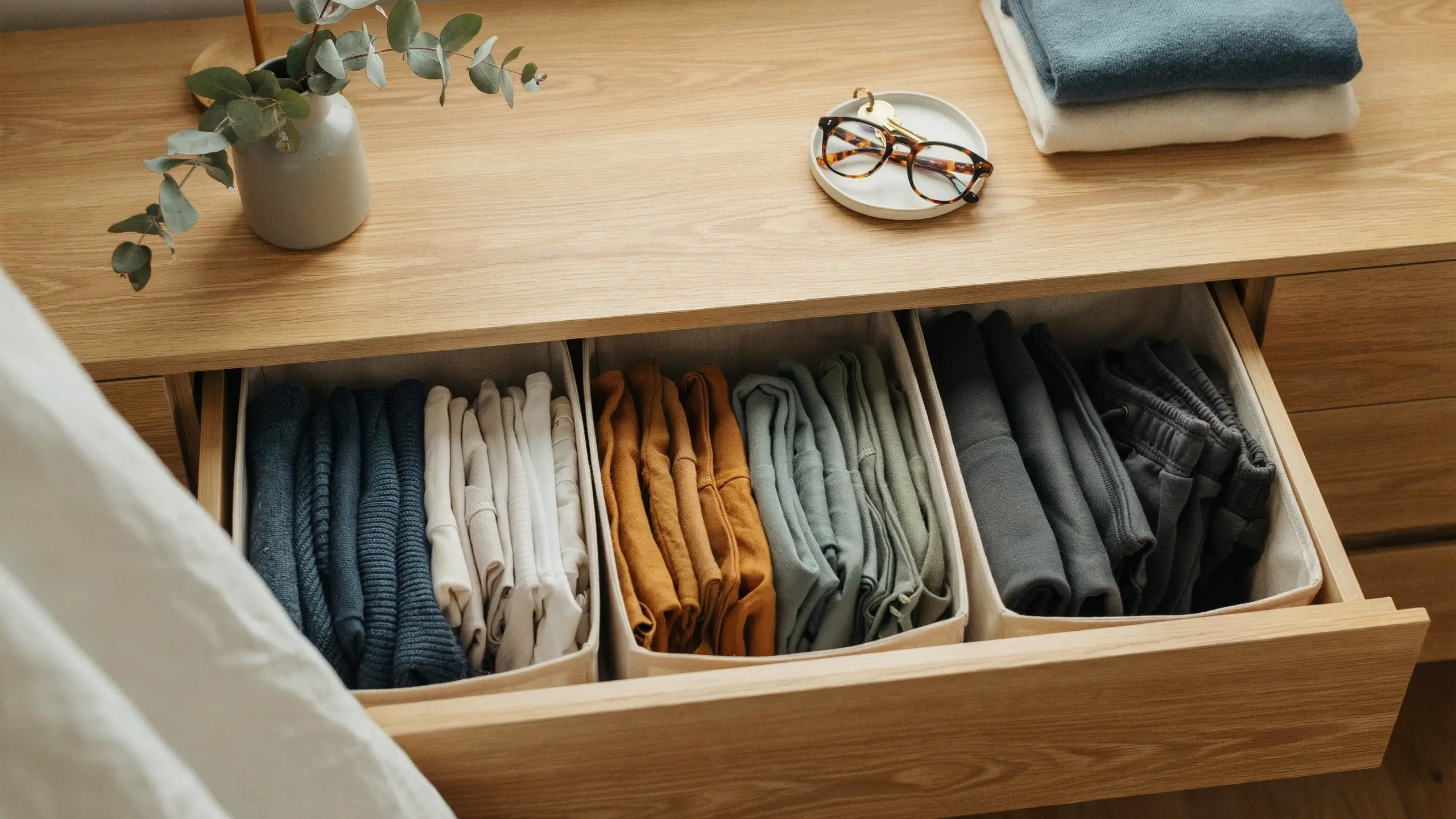 A wooden dresser drawer featuring four fabric organizers storing neatly file-folded clothes in blue, white, mustard, sage, and gray.