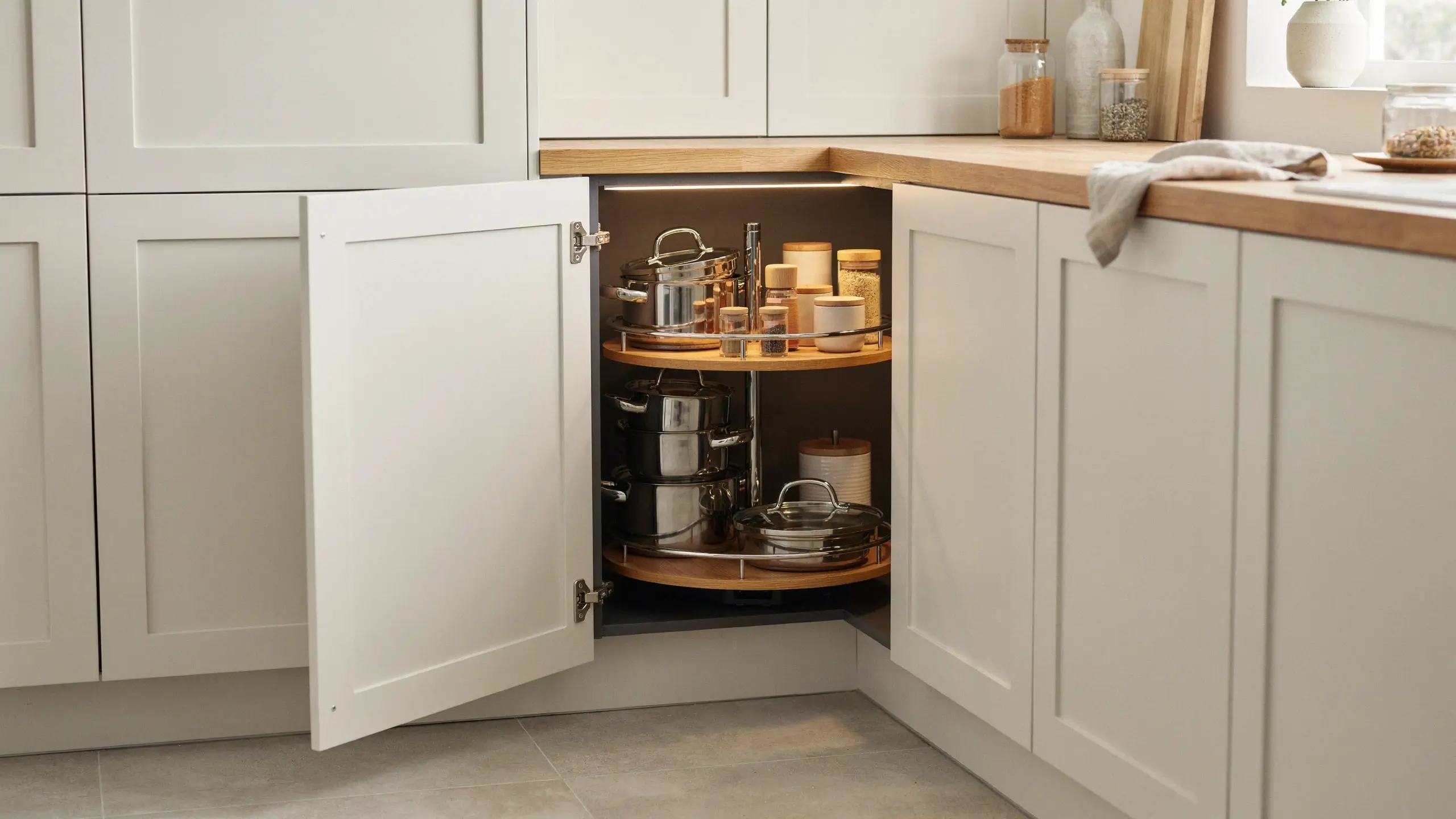 Open white shaker corner kitchen cabinet with an open door revealing a two-tier wooden lazy Susan carousel organizer filled with stainless steel cookware and clear glass spice jars.