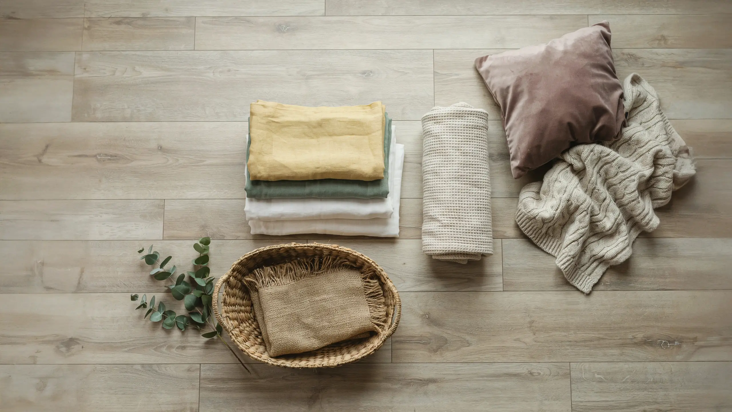 A minimalist flat lay of home textiles featuring a neatly folded stack of mustard yellow, sage green, and white linen fabrics, next to a rolled cream ribbed knit blanket, a dusty rose velvet pillow, a chunky beige cable-knit sweater, and a round wicker basket containing a brown fringed cloth and a fresh eucalyptus sprig, all arranged on a light oak wooden floor.