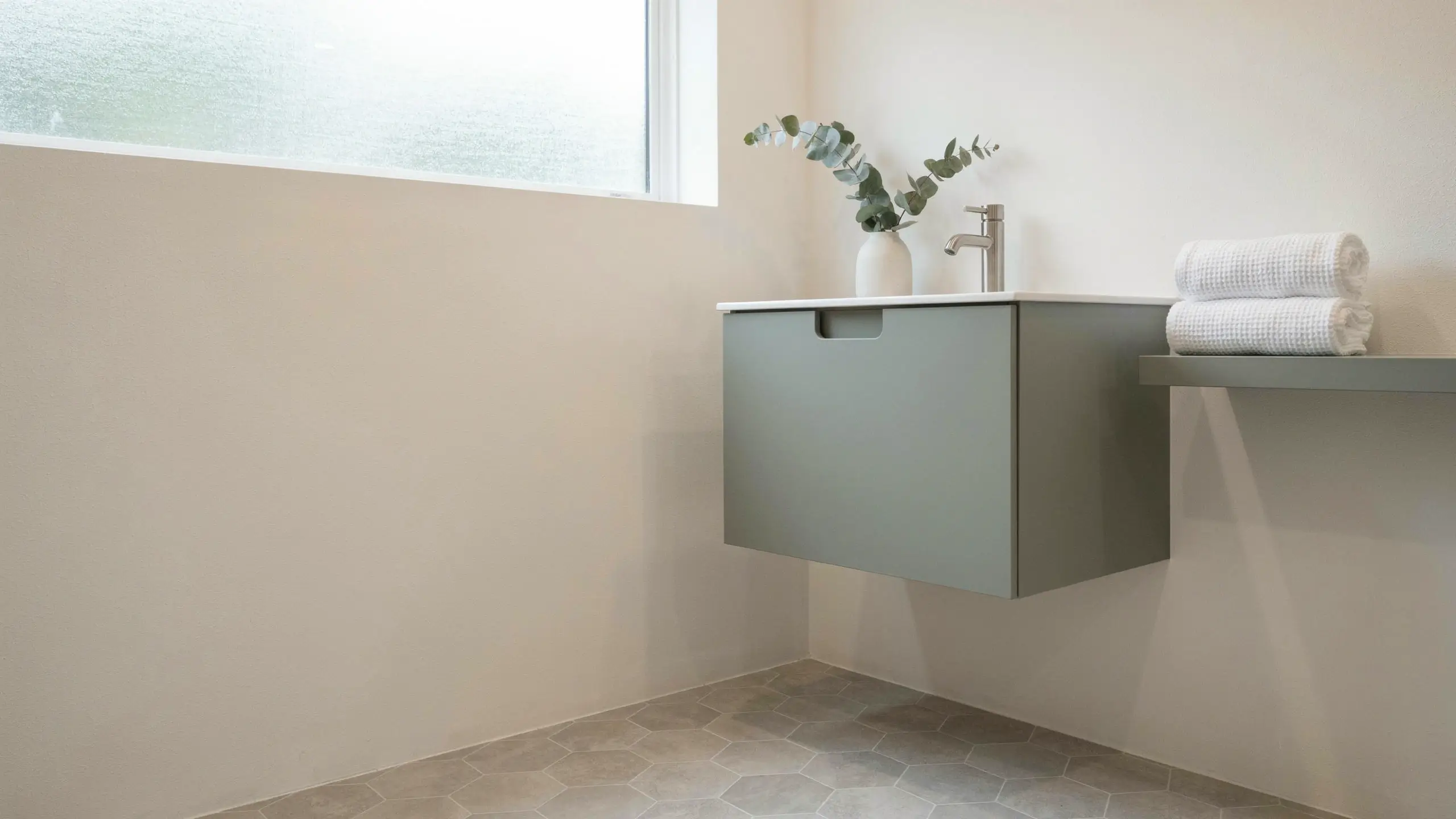 Modern floating sage green vanity with white sink and nickel faucet in a small bathroom, featuring hexagonal grey floor tiles and a white vase with eucalyptus.