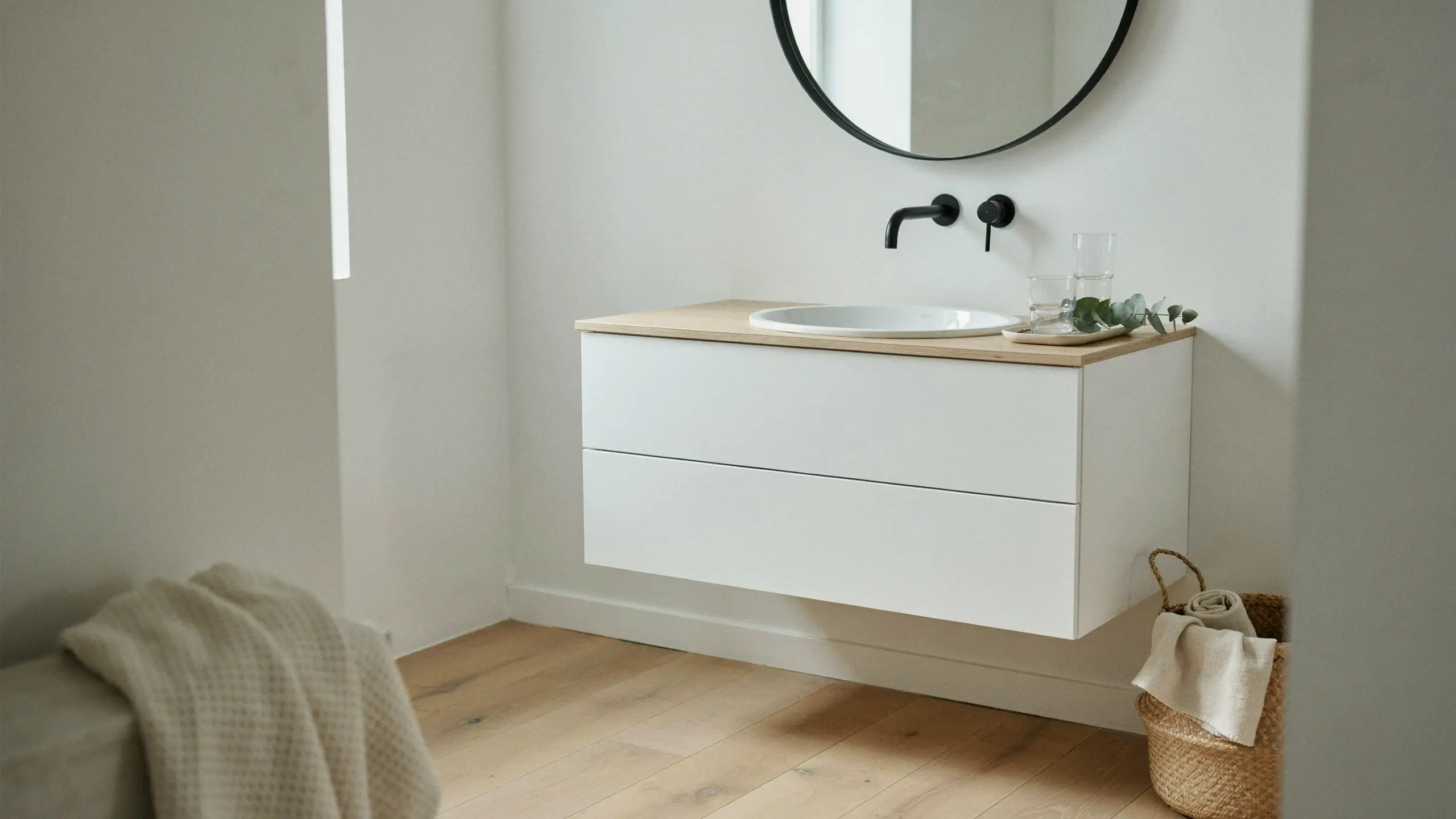 Modern floating white bathroom vanity with a light oak countertop, wall-mounted matte black faucet, and round mirror, demonstrating how to save floor space by replacing bulky cabinets.