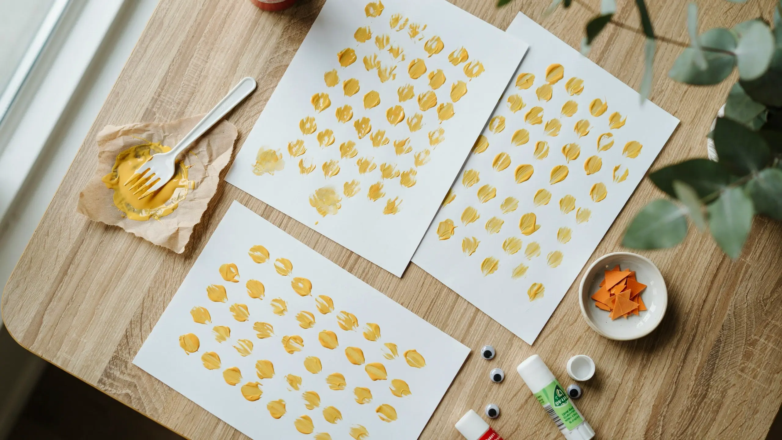 Overhead flat lay of a craft table setting for making fork-painted chicks. White paper displays rows of yellow paint stampings created with a plastic fork, surrounded by googly eyes, a bowl of small orange paper triangles for beaks, and a glue stick for assembling the DIY spring art project.