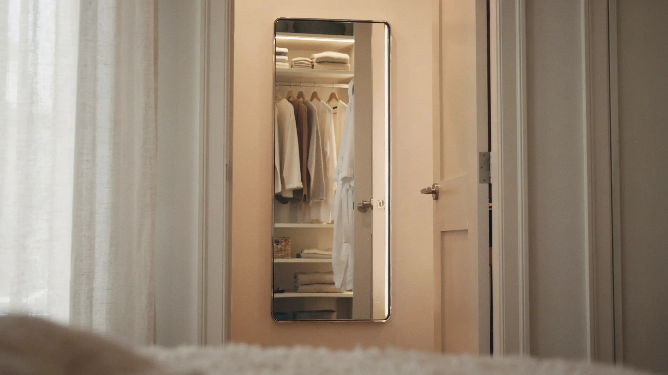 Slim full-length rectangular mirror with a thin metal frame mounted on a white bedroom door, clearly reflecting a neatly organized walk-in closet with hanging neutral-toned garments, a white robe, wooden shelving, and stacked folded towels