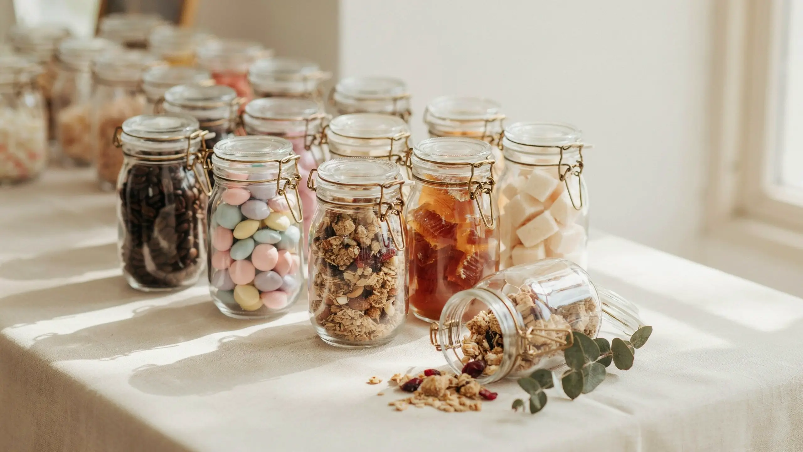A row of glass airtight storage jars with wire bail latches holding assorted pantry snacks, including dark chocolate chips, pastel candy-coated chocolates, granola with dried cranberries, sliced apricots, and marshmallows. In the foreground, a tipped jar spills granola over a white linen tablecloth beside a sprig of fresh eucalyptus leaves.