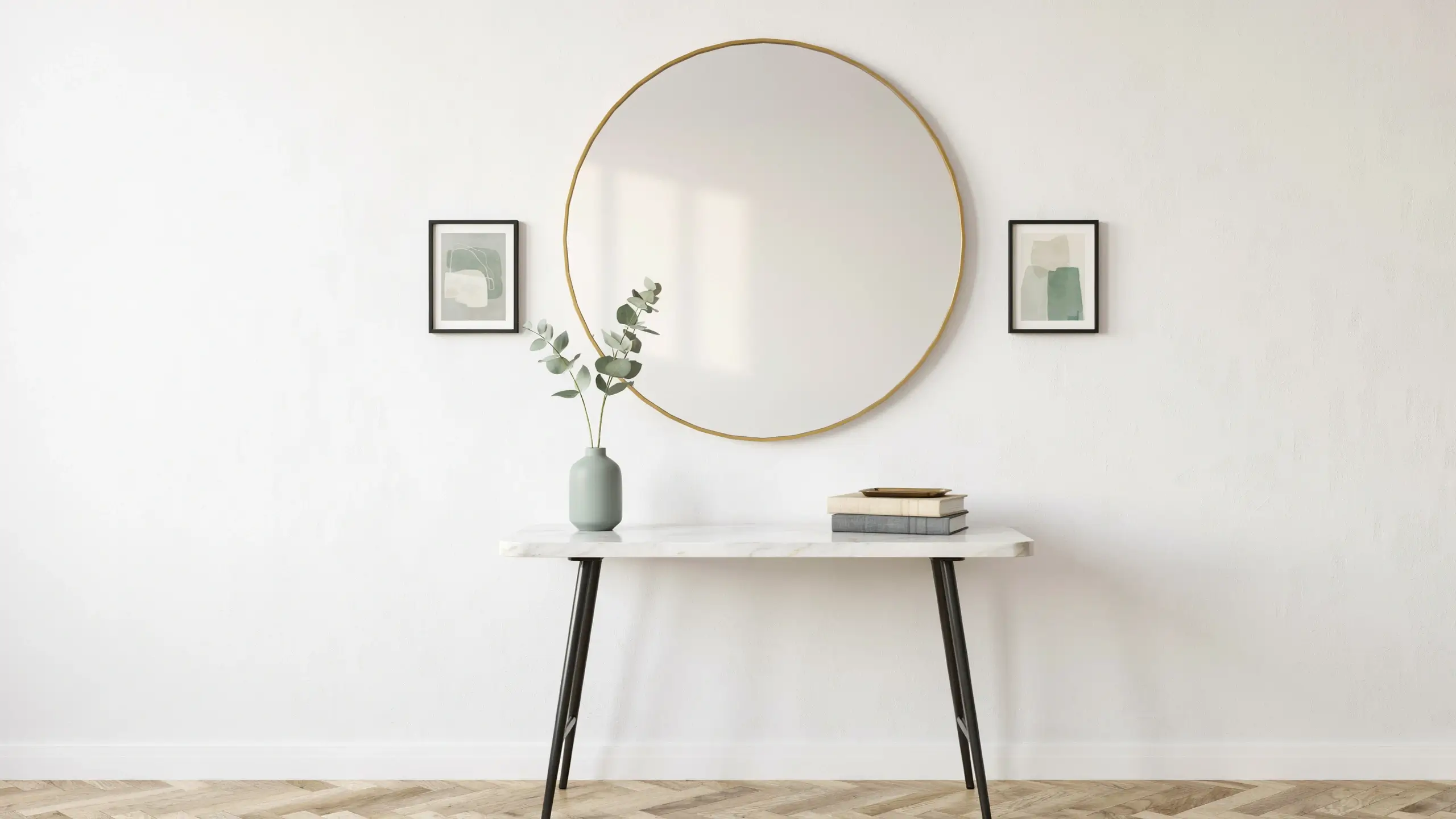 Large round gold-framed mirror serving as a wall focal point in a minimalist modern entryway, styled with a white marble console table, sage green ceramic vase containing eucalyptus stems, neutral abstract framed wall art, and a stack of decorative books.