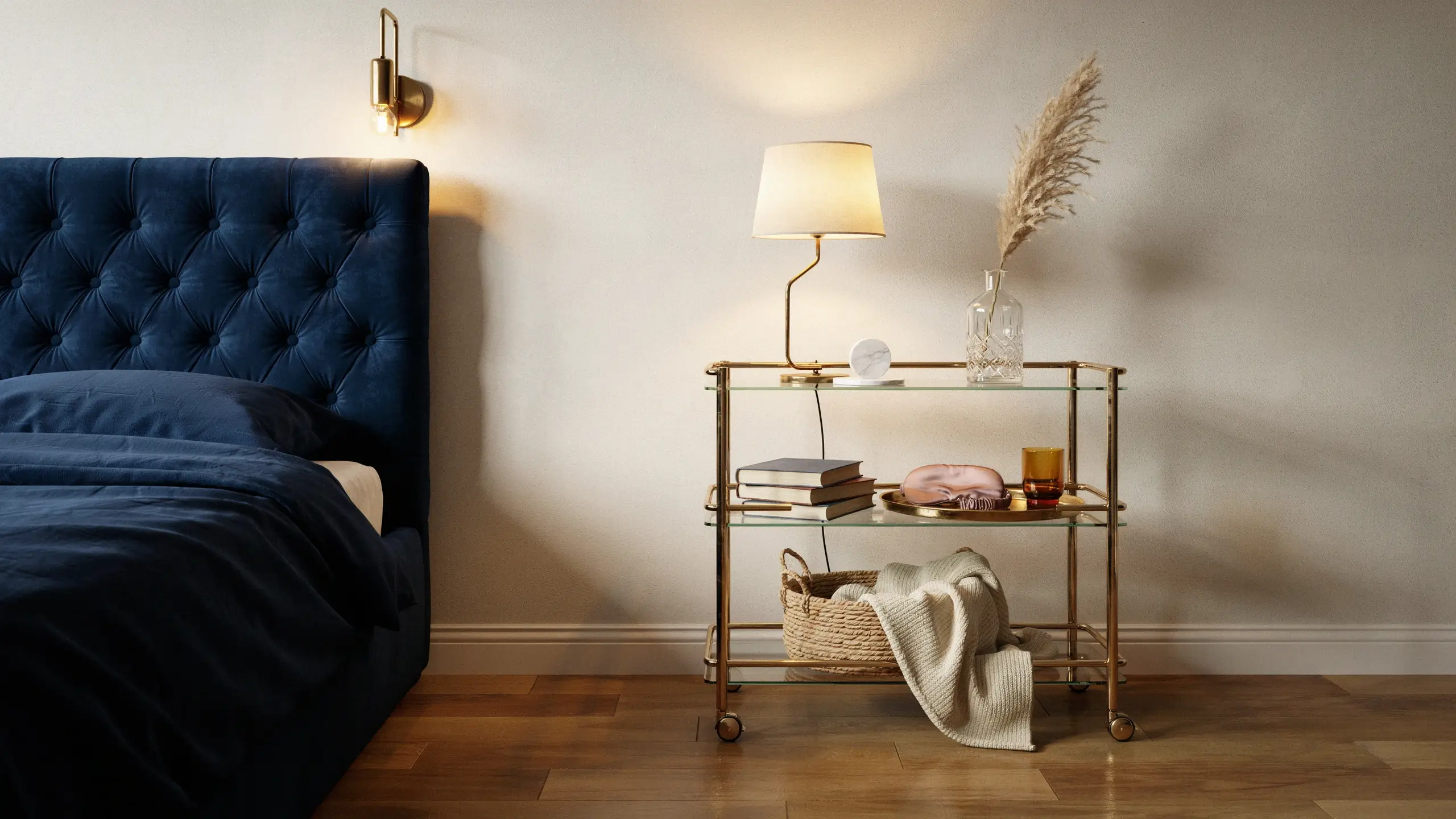 A stylish gold-framed glass rolling bar cart styled as a bedside drinks station, holding a brass table lamp, a glass vase with pampas grass, and books, situated next to a plush navy blue tufted bed in a cozy, elegant bedroom. 