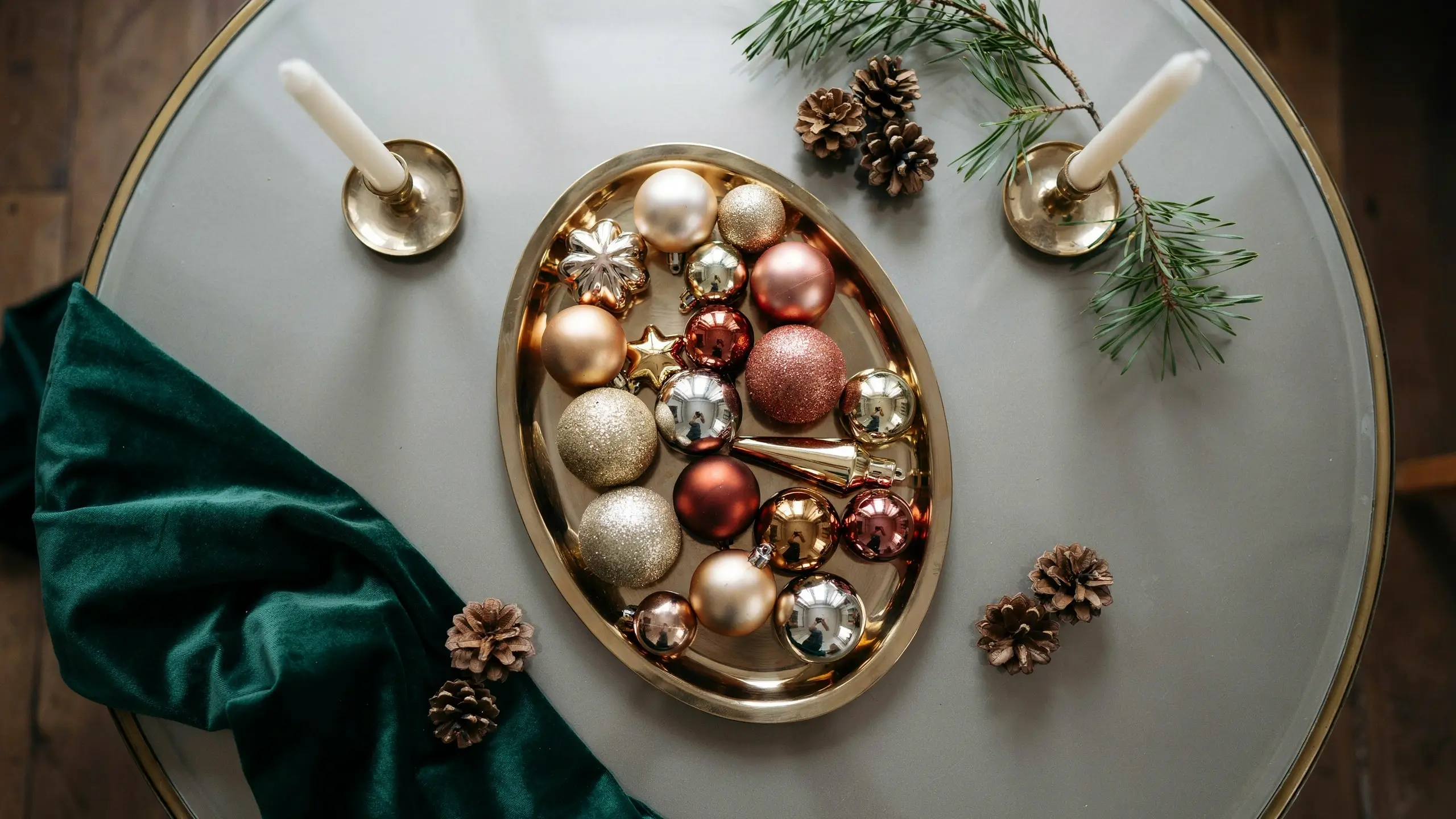 An elegant top-down view of a festive centerpiece featuring a gold oval tray filled with mixed metallic rose gold, silver, and gold Christmas ornaments, accented with white taper candles, pine sprigs, and a draped dark green velvet fabric.