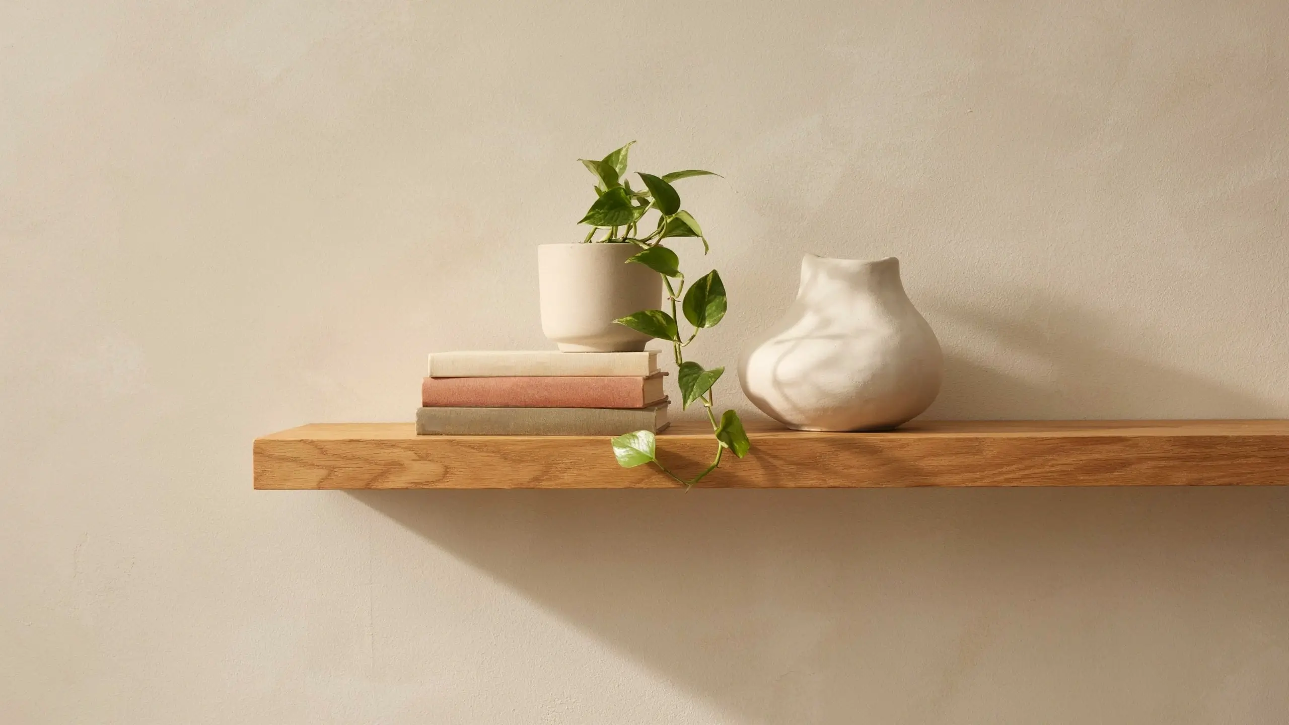 A minimalist grouping of decor on a floating shelf featuring a stack of three earthy-toned books, a white potted trailing pothos plant, and a sculptural ceramic vase against a textured light beige wall.