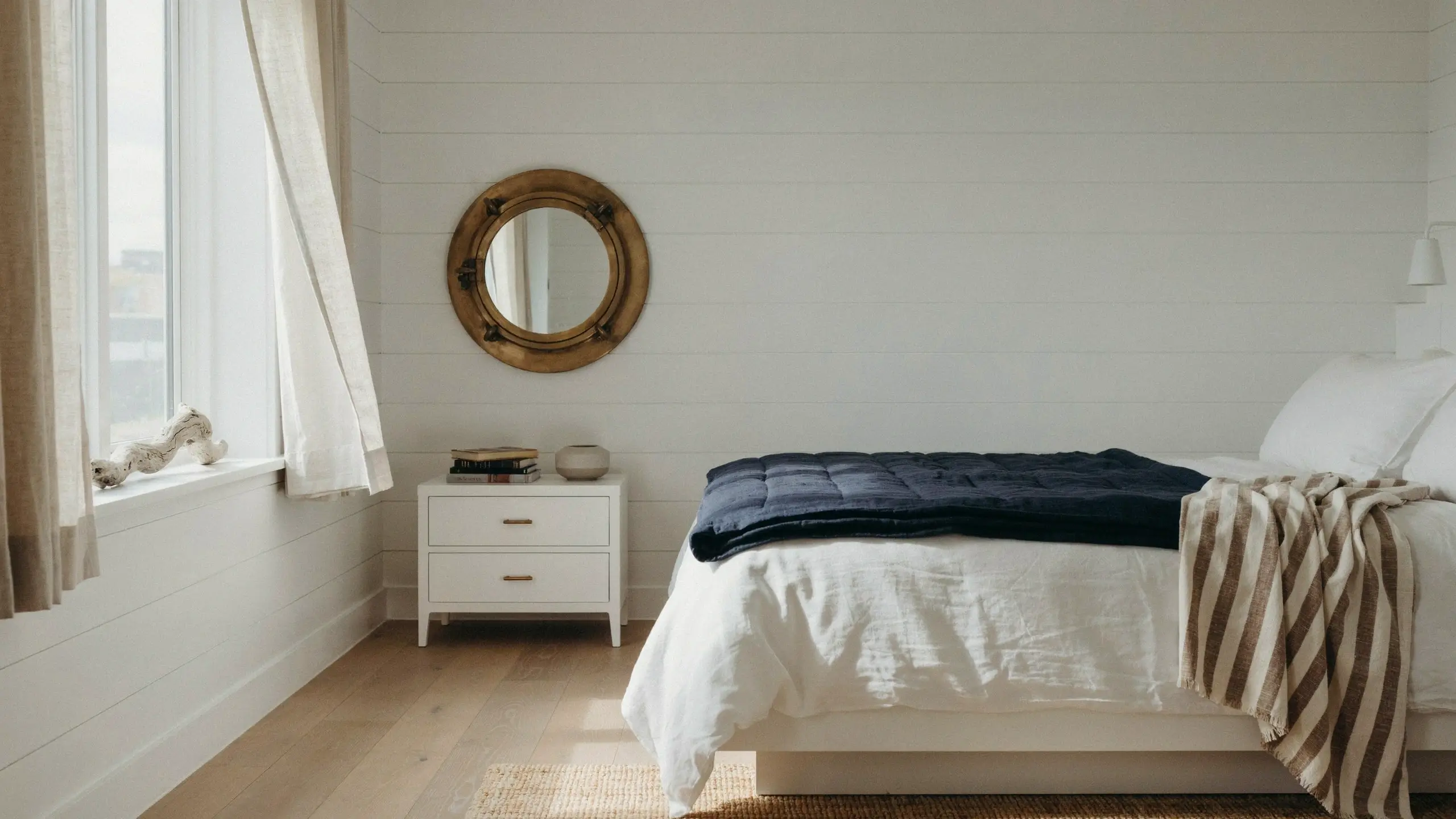 A bright and serene grown-up coastal nautical bedroom featuring white shiplap walls, a circular brass porthole mirror, a white nightstand, and a bed layered with white linens, a navy quilted throw, and a striped beige blanket.