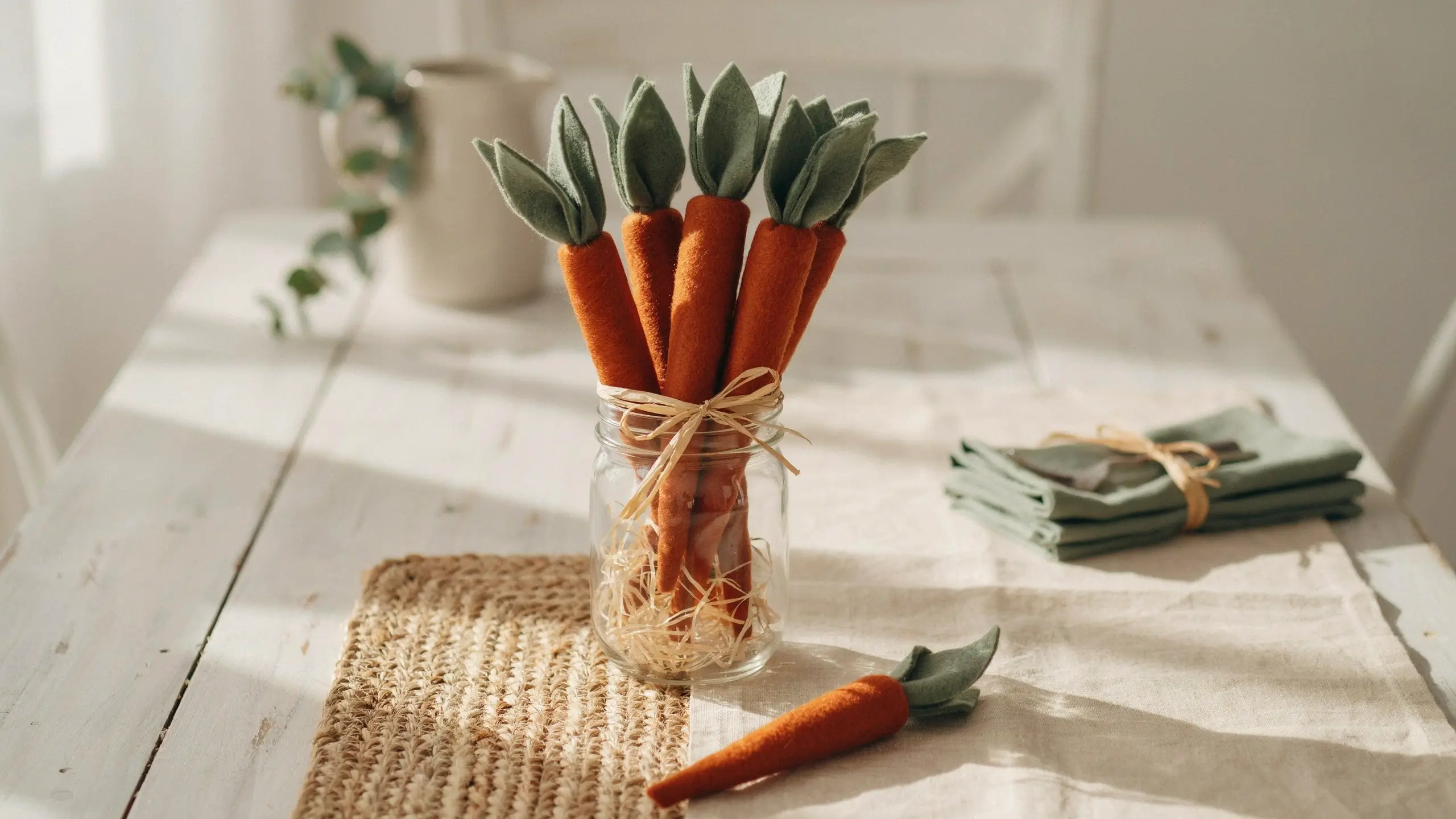Handmade felt carrot bundle centerpiece in a clear glass mason jar tied with rustic jute twine, sitting on a white wooden table with a woven placemat, sage green napkins, and a loose carrot in the foreground.