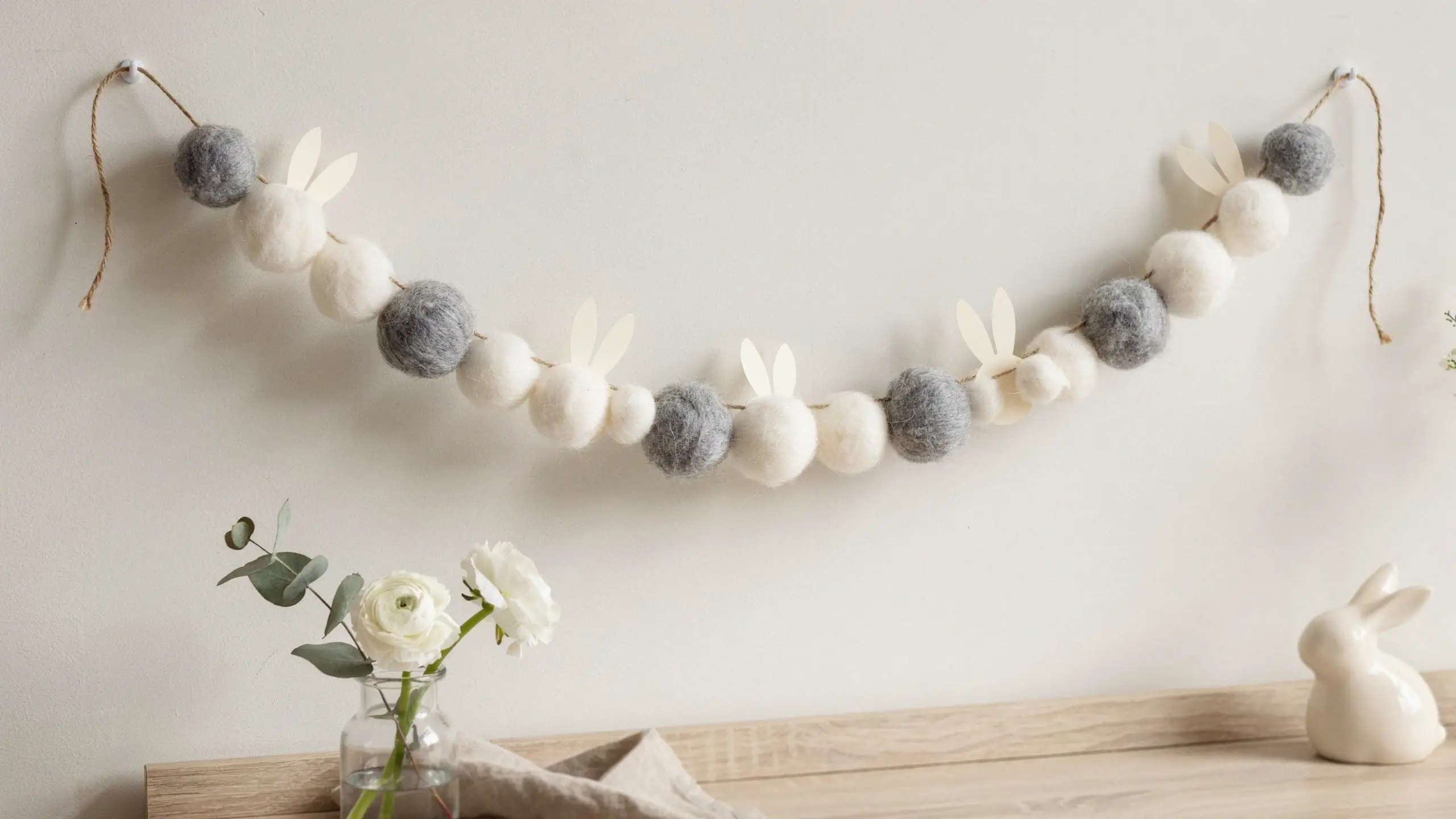 Handmade white and grey felt pom-pom bunny garland hanging on a wall with twine for spring Easter nursery decor.