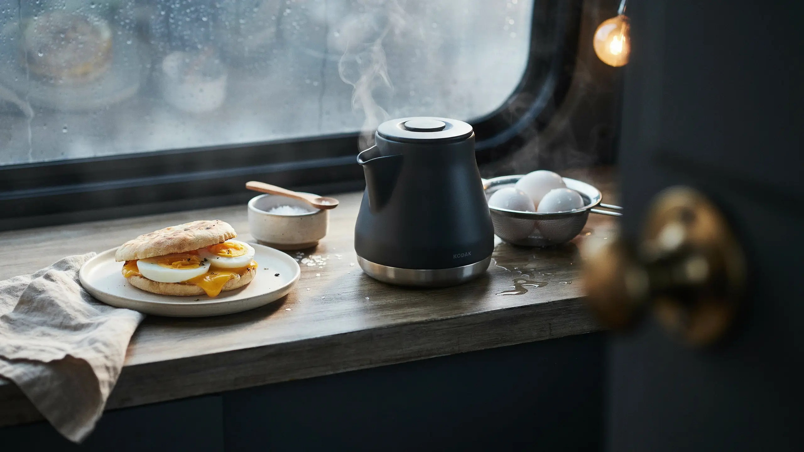 Hot breakfast sandwich with kettle-boiled eggs and melted cheese placed on a rustic wooden counter next to a steaming black electric kettle and a rainy window.