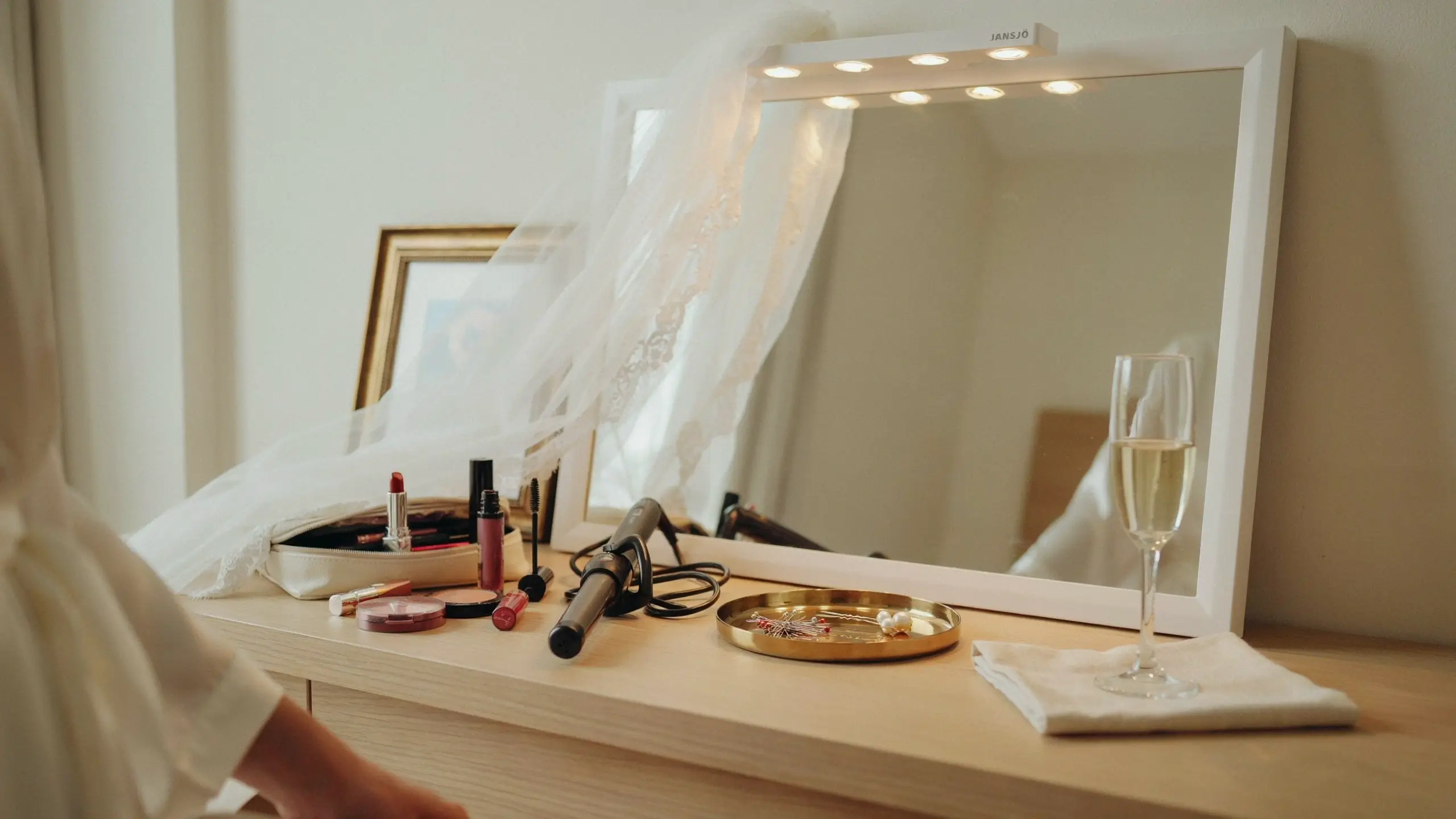 An IKEA JANSJÖ LED clip light attached to a white vanity mirror, illuminating a bridal getting-ready setup with a white lace veil, makeup bag, cosmetics, curling iron, and a champagne flute.