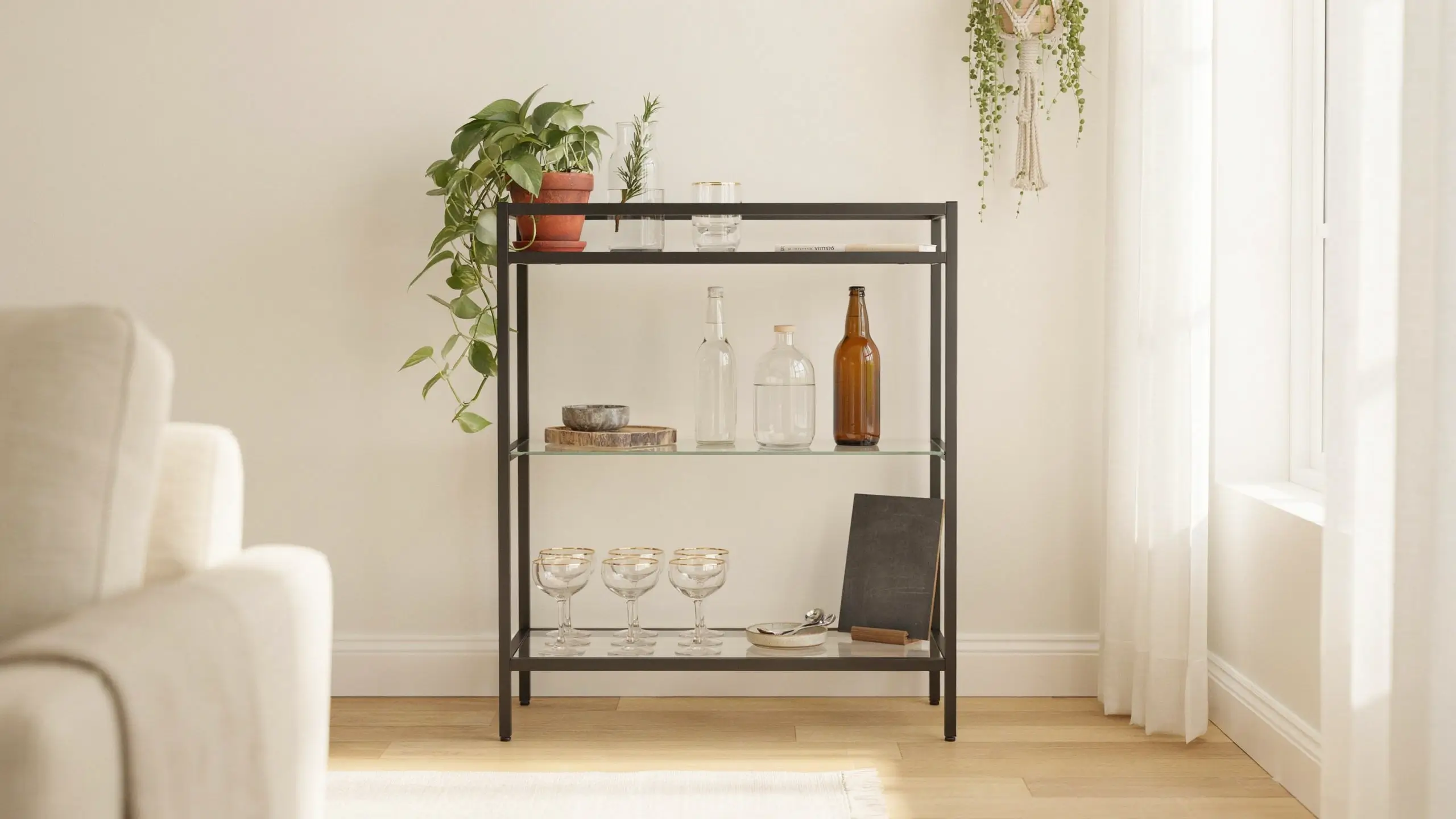 An IKEA VITTSJÖ black metal and glass shelving unit repurposed as a minimalist bar cart. The home bar setup features glass bottles, gold-rimmed stemware, a trailing pothos plant, and a chalkboard sign, styled in a bright airy living room with white walls and sheer curtains.