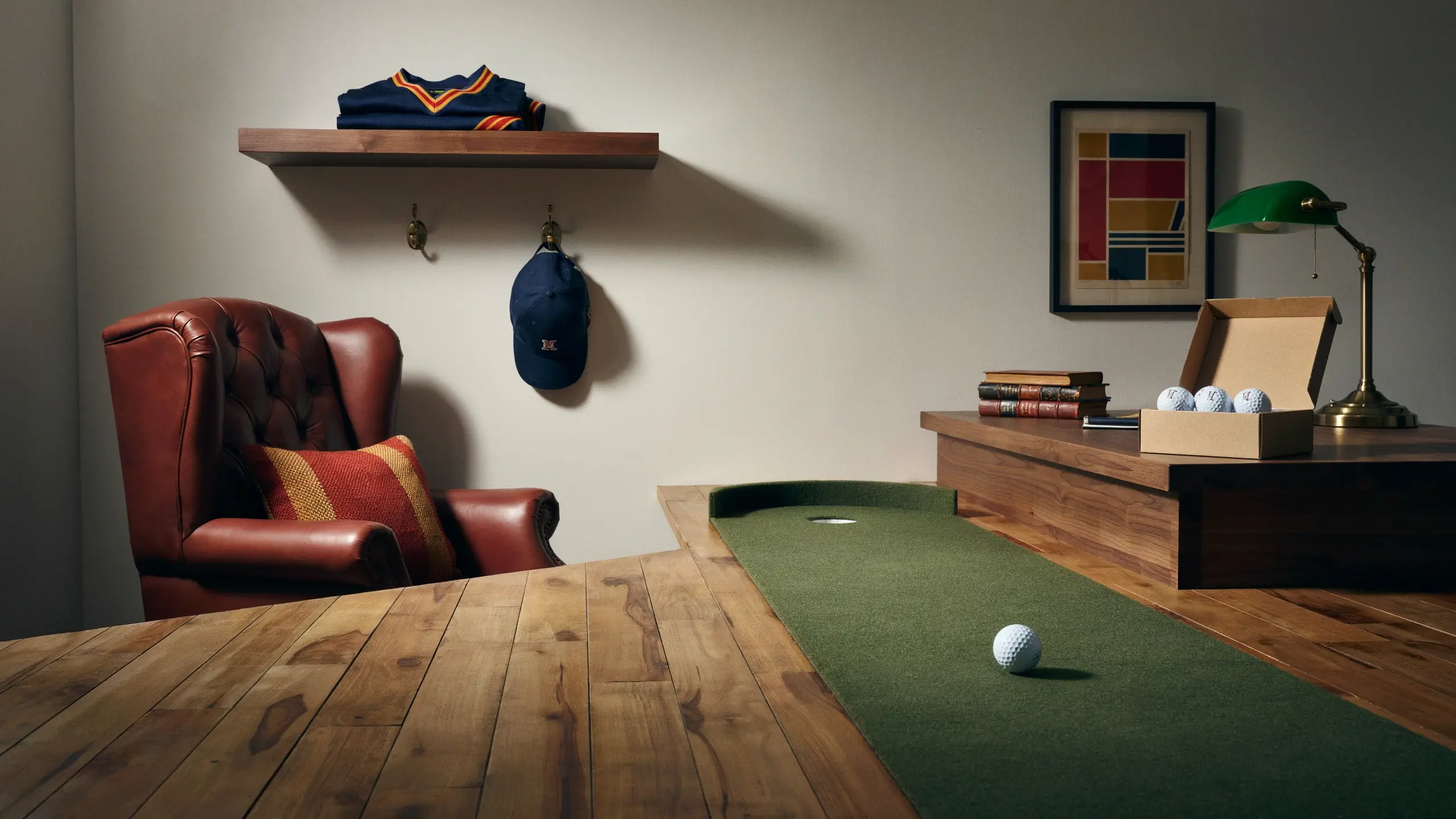A cozy indoor sports den with a wooden floor featuring a golf putting green and a white golf ball. To the left sits a red leather tufted wingback armchair with a striped pillow, while a blue cap and folded athletic shirt hang on a mounted wooden shelf. On the right, a desk holds a green banker's lamp, stacked books, and an open box of white golf balls. 