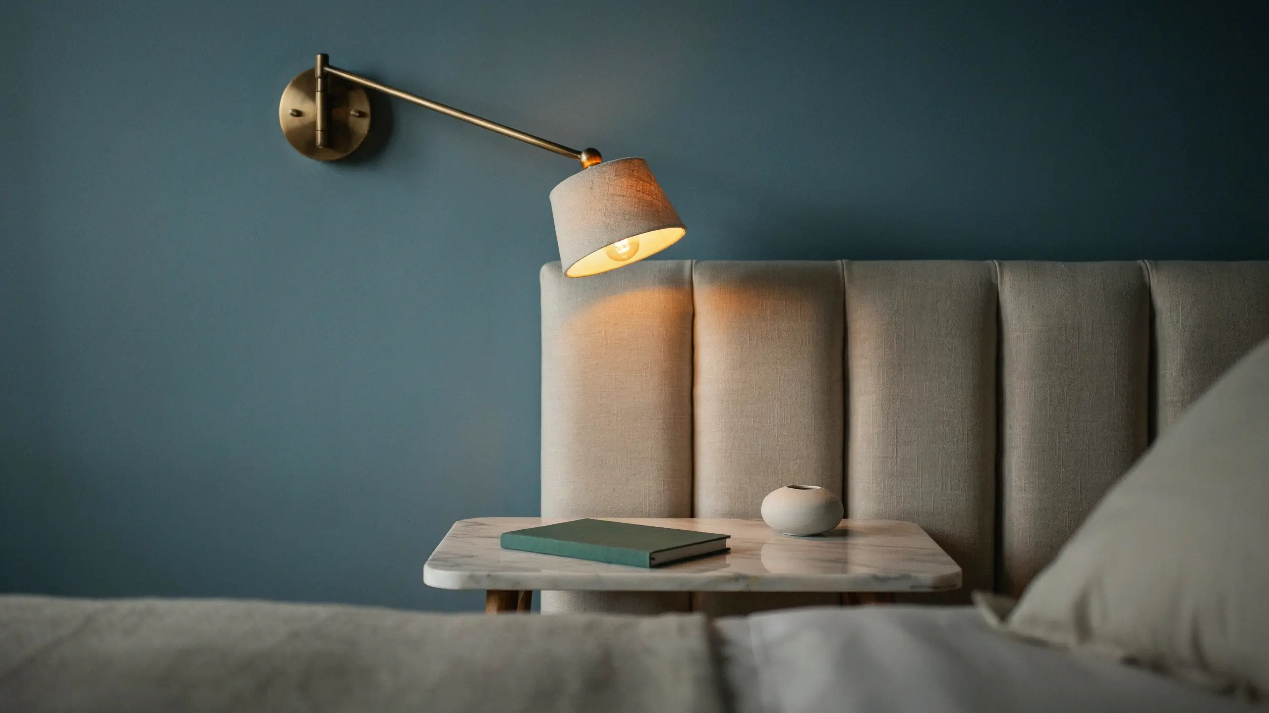 A modern wall-mounted swing arm sconce with a beige fabric shade projecting warm reading light over a marble bedside table with a green book and white ceramic decor, providing a space-saving alternative to bulky table lamps in a contemporary bedroom.