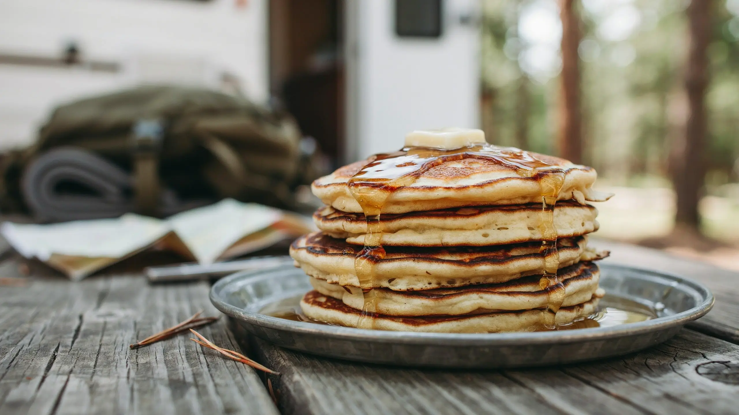 Fluffy stack of Kodiak Cakes protein pancakes topped with melting butter and maple syrup on a metal camping plate outdoors.