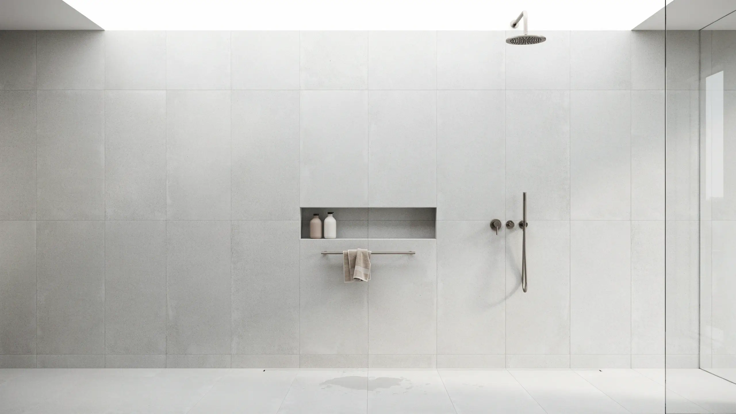 Minimalist walk-in shower featuring large-format light grey wall tiles with nearly invisible grout lines, complete with a rainfall showerhead, wall-mounted controls, and a recessed niche storing bathroom bottles.